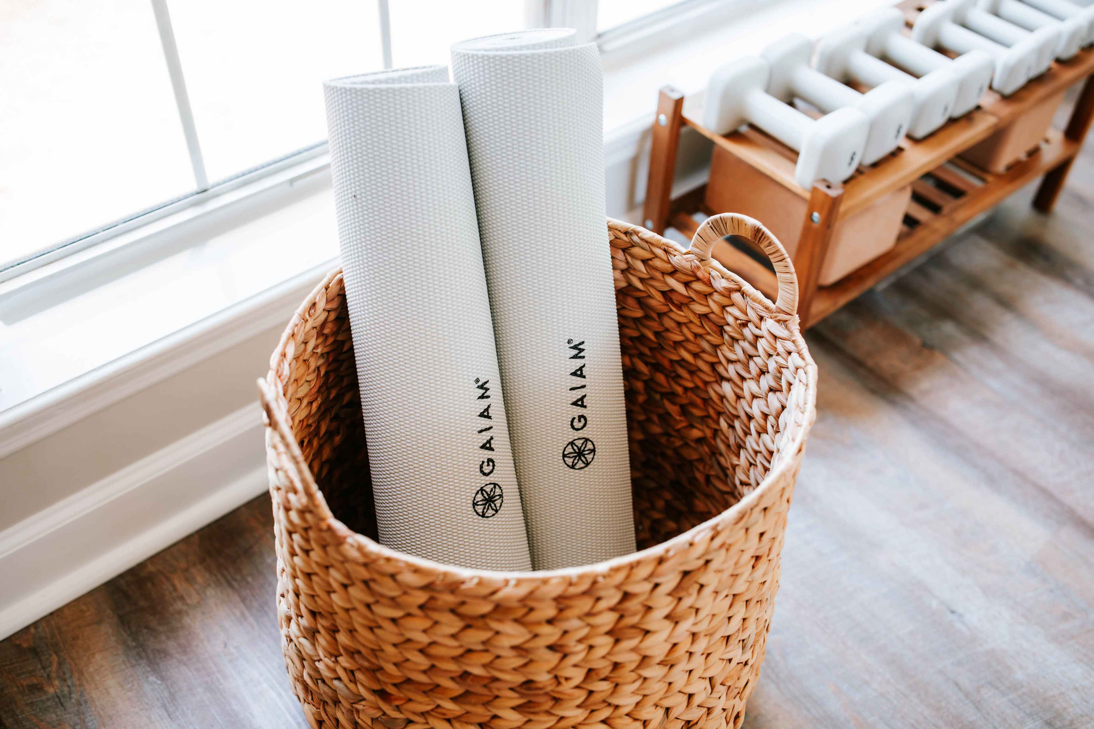 Two yoga mats are stored upright in a woven basket beside a wooden rack of dumbbells.