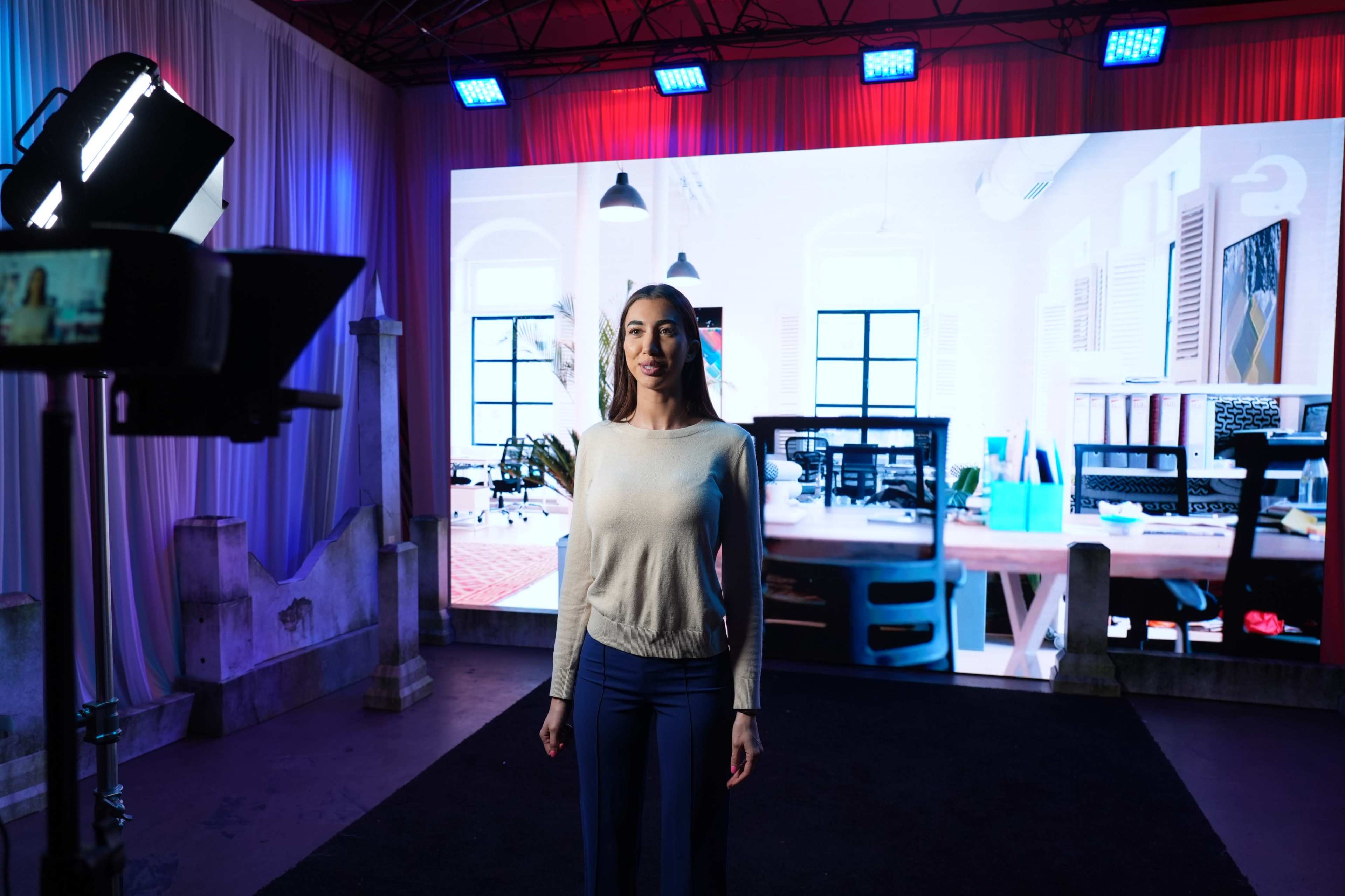 A woman stands in front of a large screen displaying a modern office setting, illuminated by overhead lights in a studio environment.