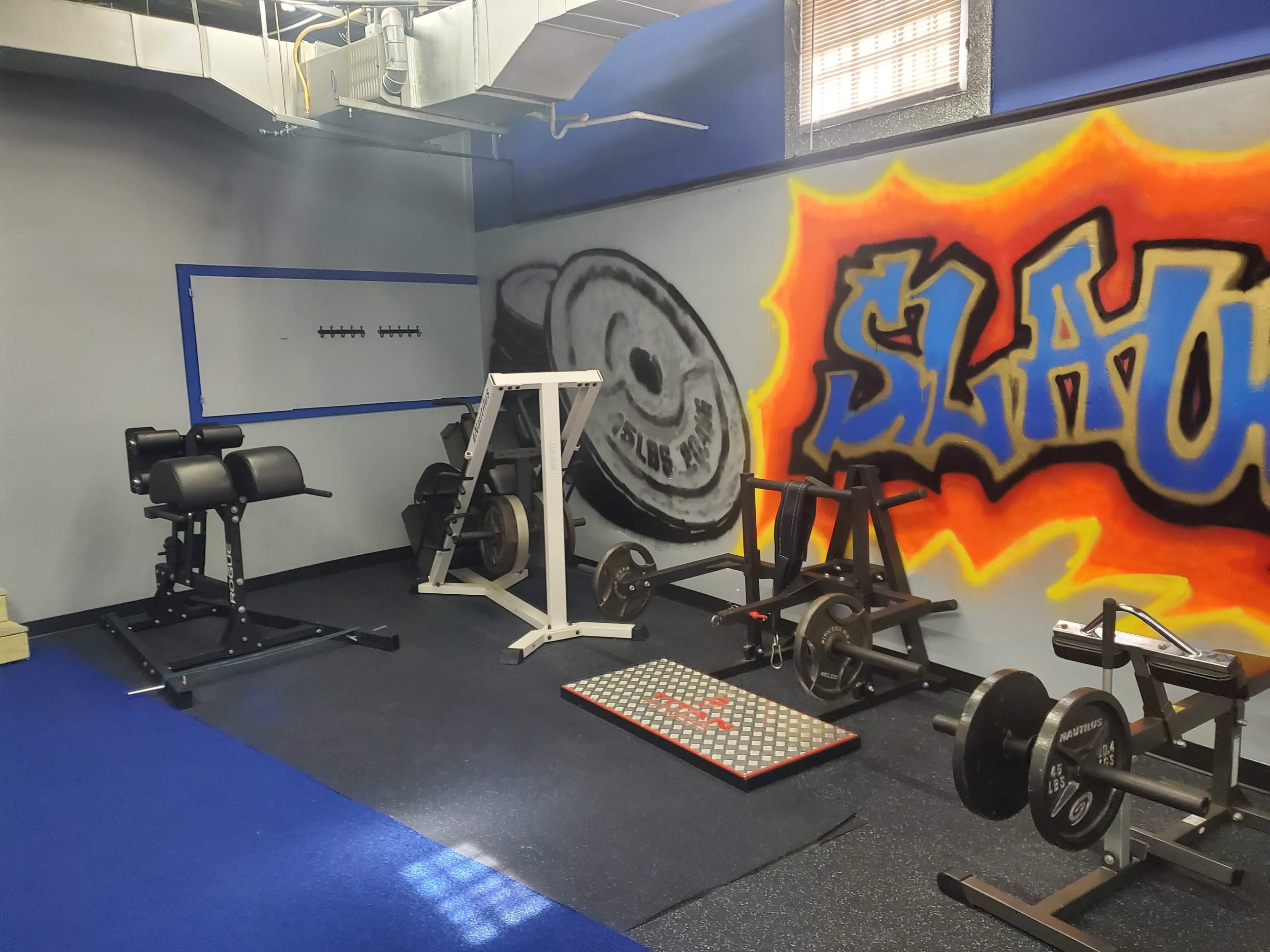 A small gym space equipped with various weightlifting equipment, a foam exercise mat, and a colorful graffiti-style mural on the wall.