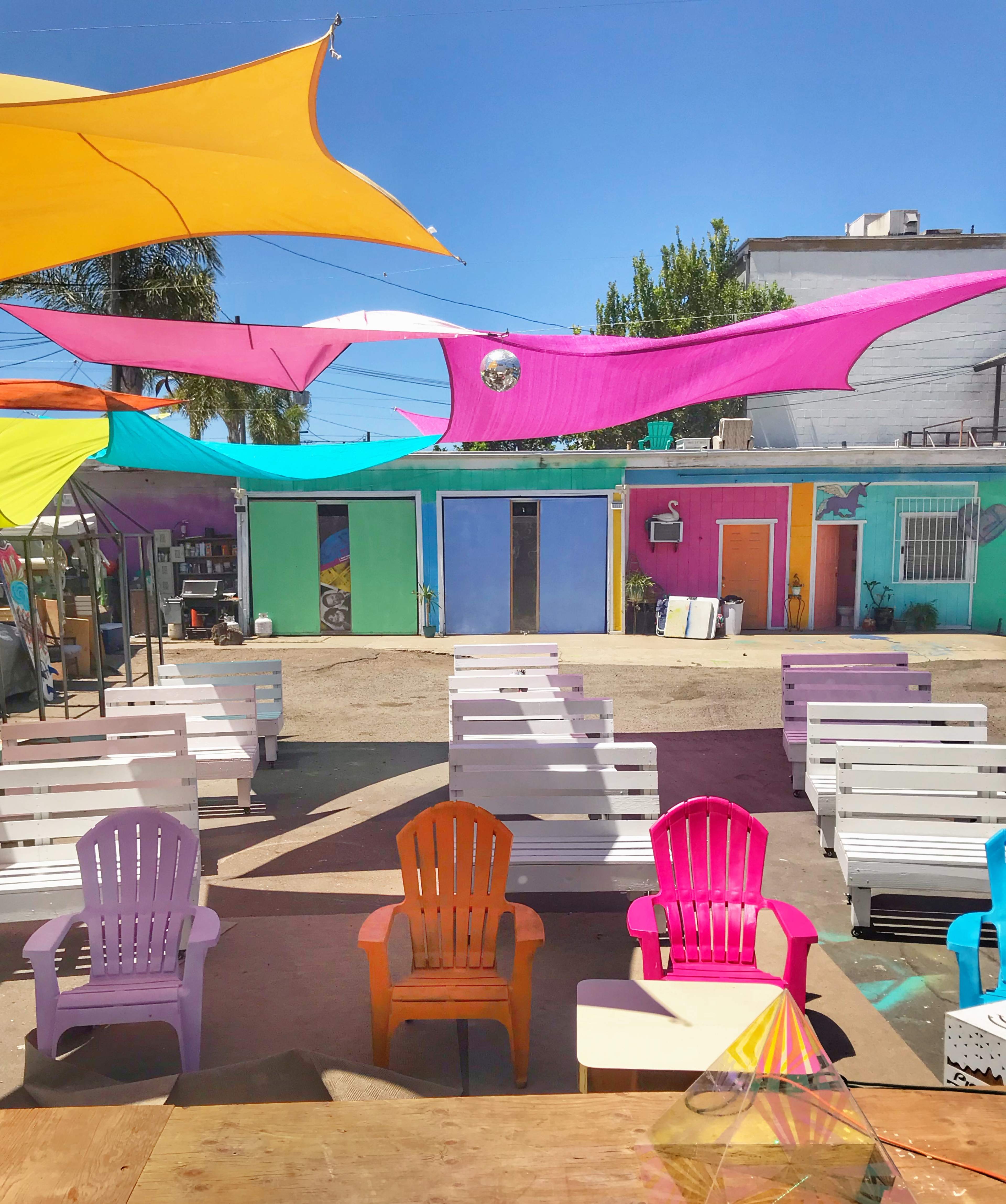 A vibrant outdoor space features colorful canopies overhead and a cluster of white lounge chairs arranged in front of a row of brightly painted buildings.