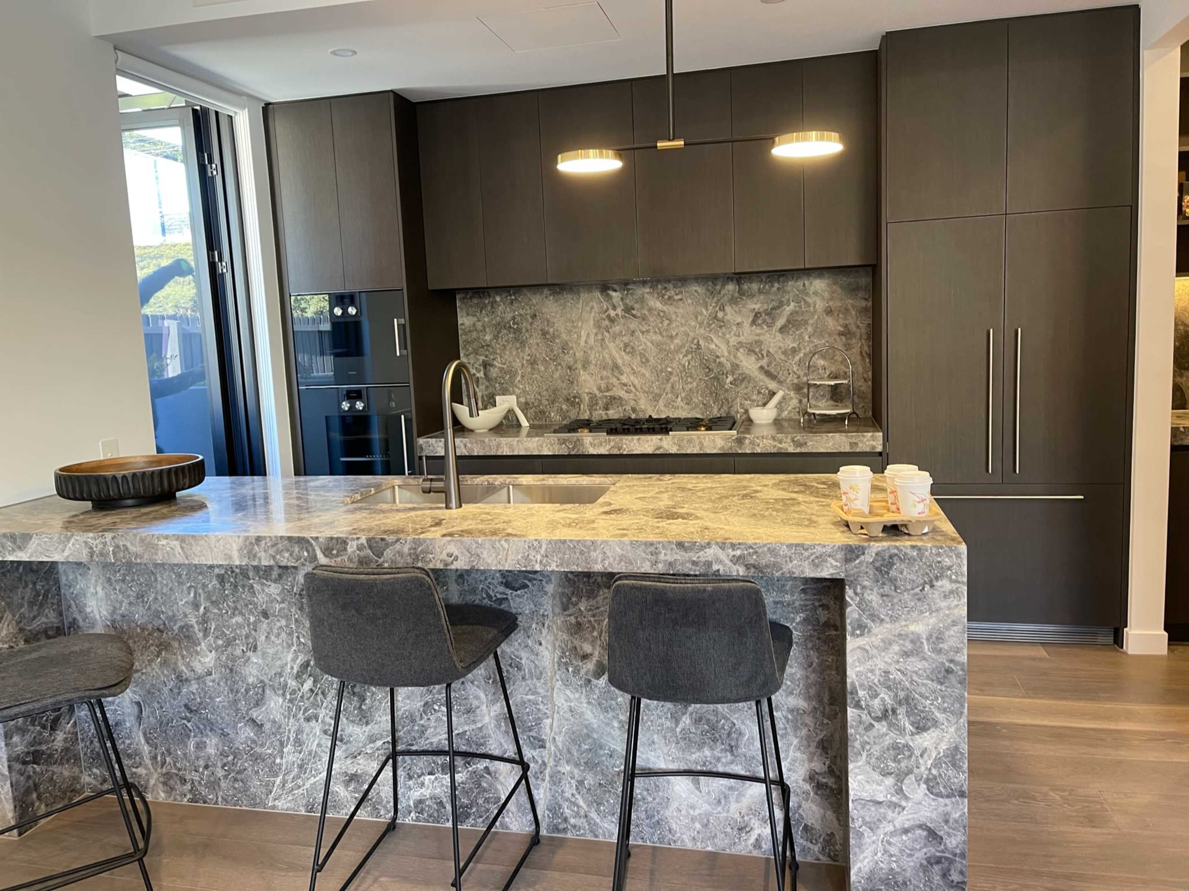 A modern kitchen features a dark cabinetry design, a marble countertop, and bar stools at an island.