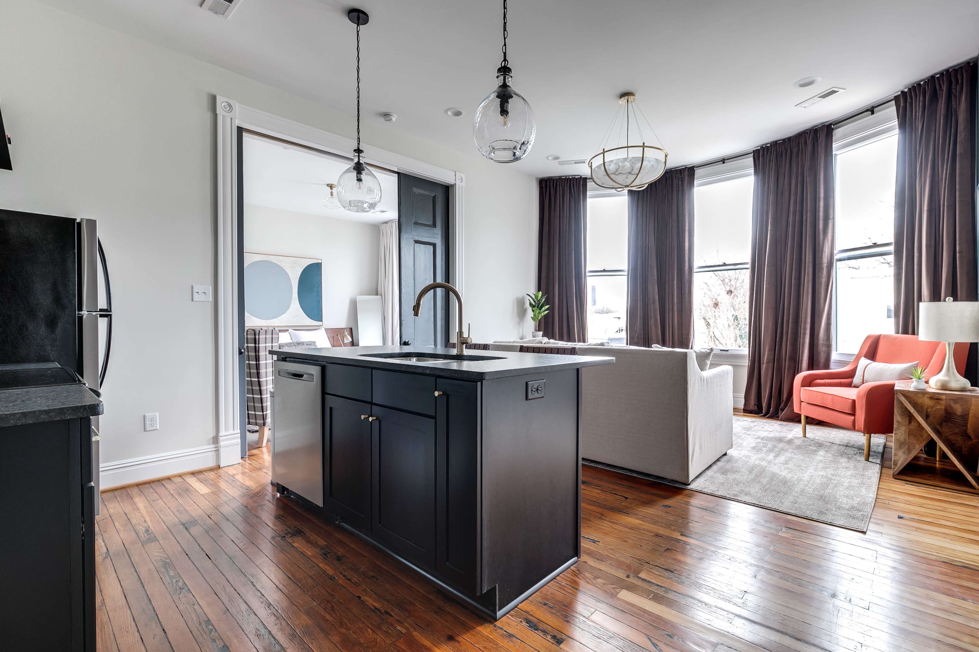 A modern kitchen features a central island with a dark finish, leading to an open living area with large windows and stylish furnishings.