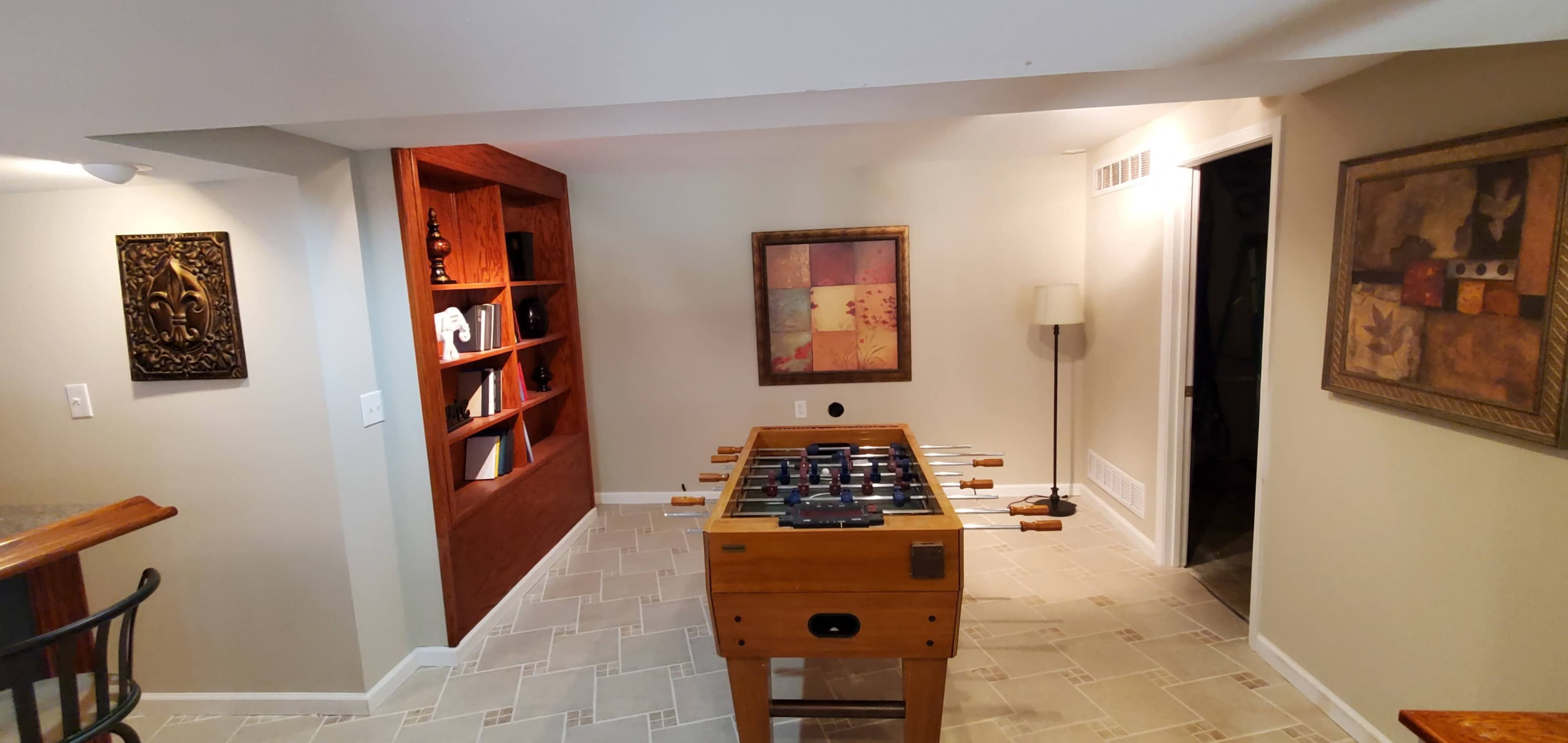 A room with a foosball table in the center, surrounded by shelves and a wall art piece, along with a lamp in the corner.