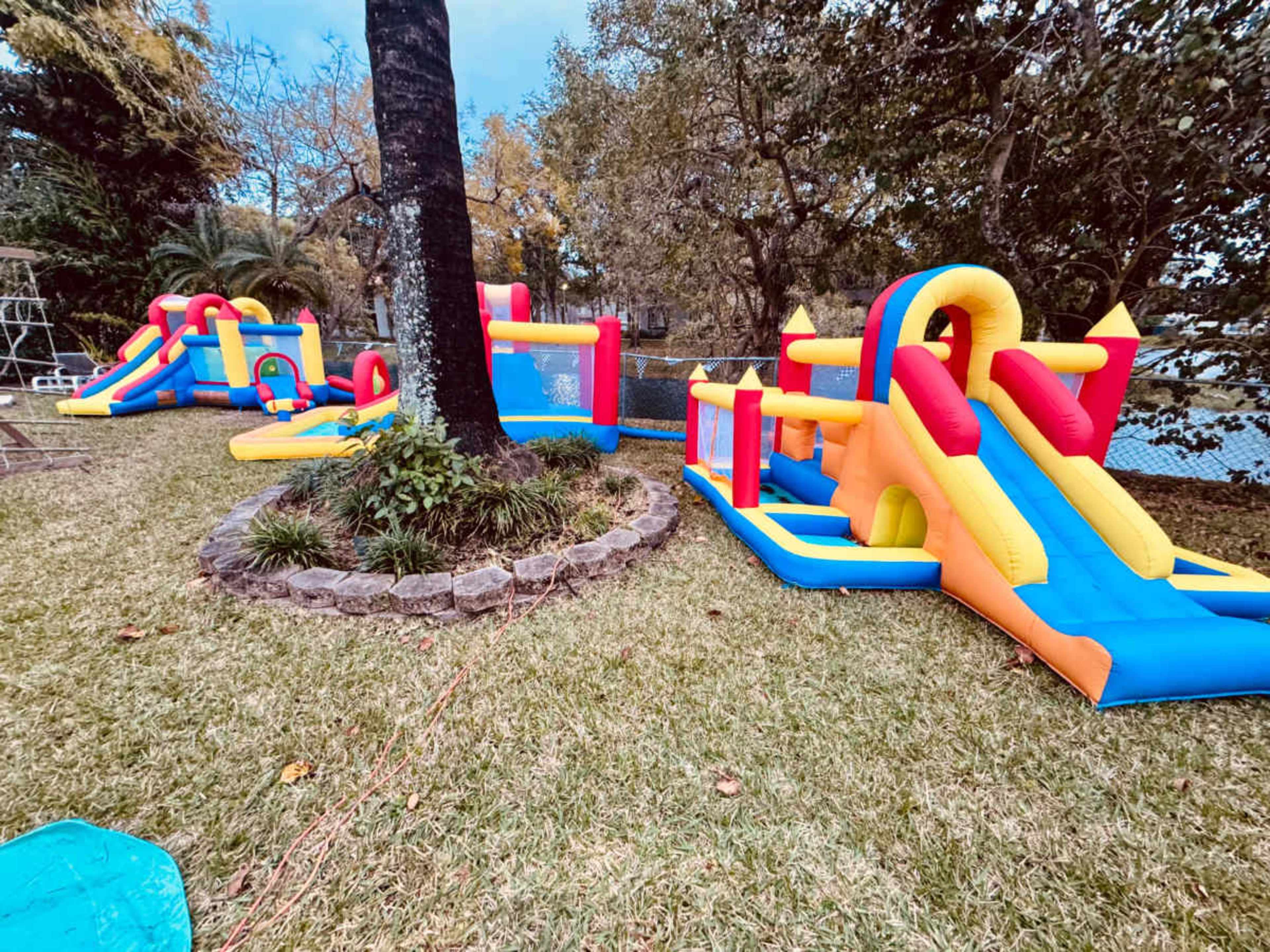 The image shows a colorful inflatable bounce house and slide setup in a grassy backyard near a pool.