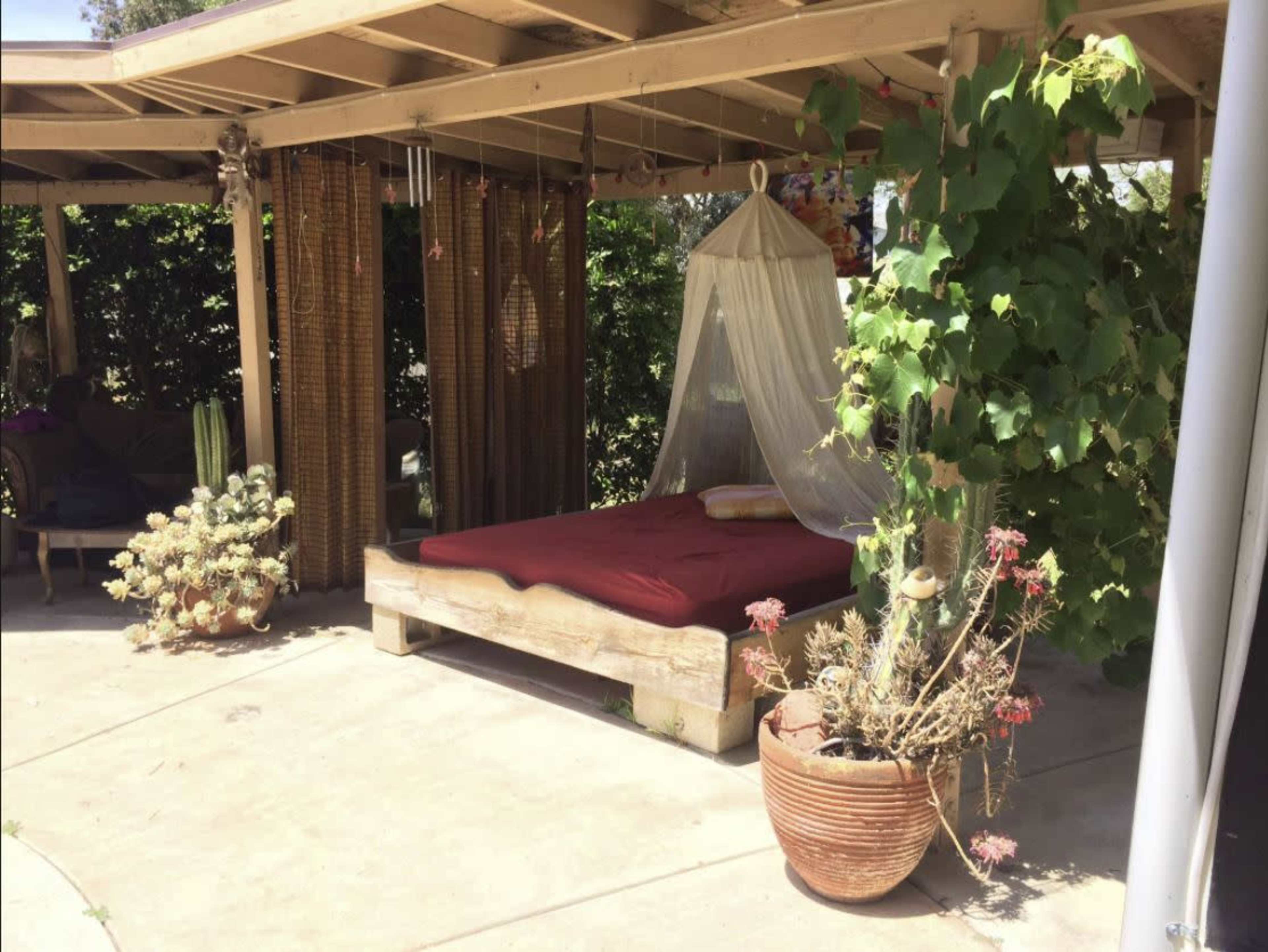 The image shows a shaded outdoor space featuring a wooden bed with a maroon mattress, surrounded by potted plants and hanging curtains.