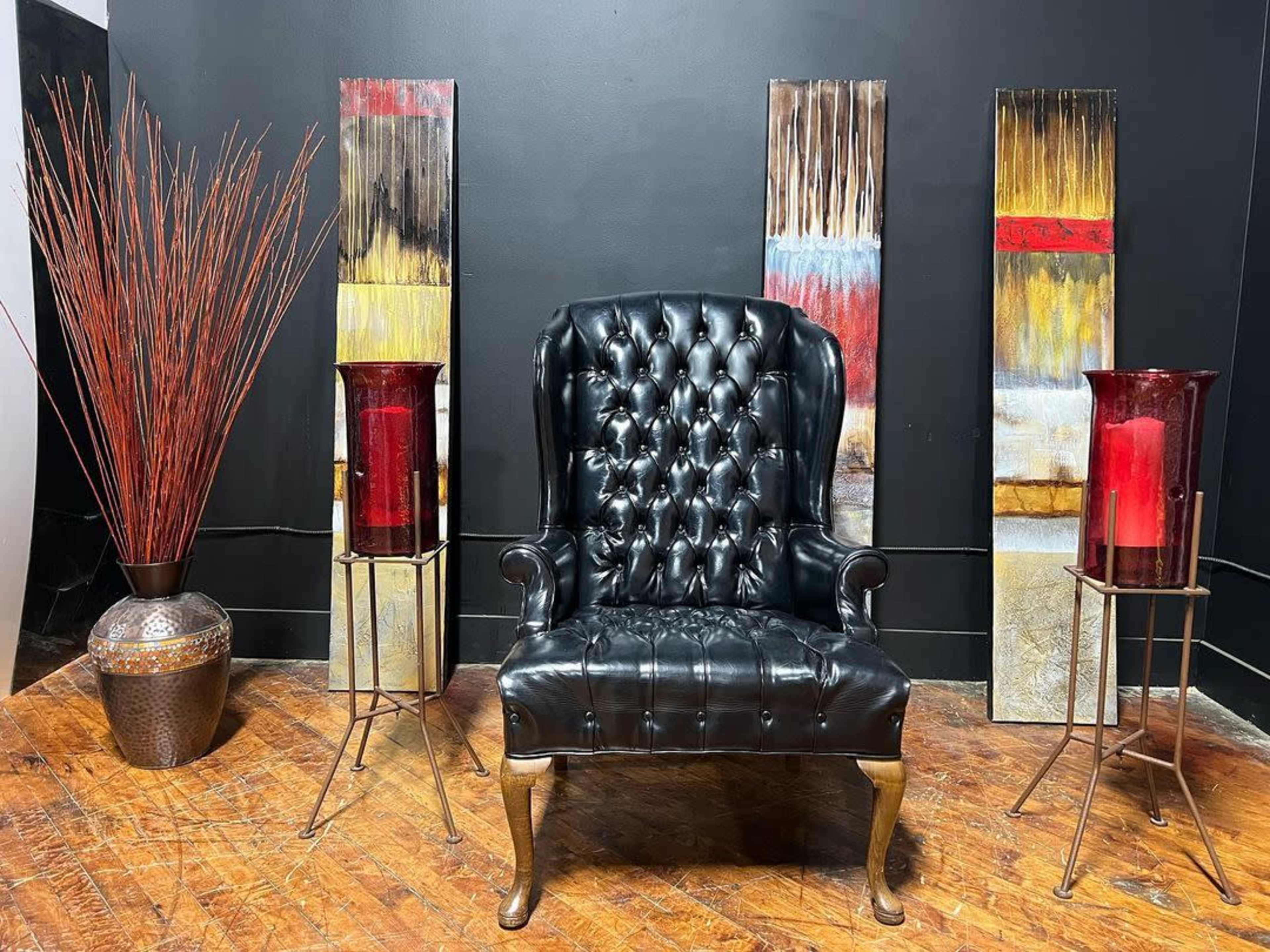 The image features a black tufted armchair placed in front of two abstract art panels, with tall decorative vases and a large pot of ornamental grass on either side.