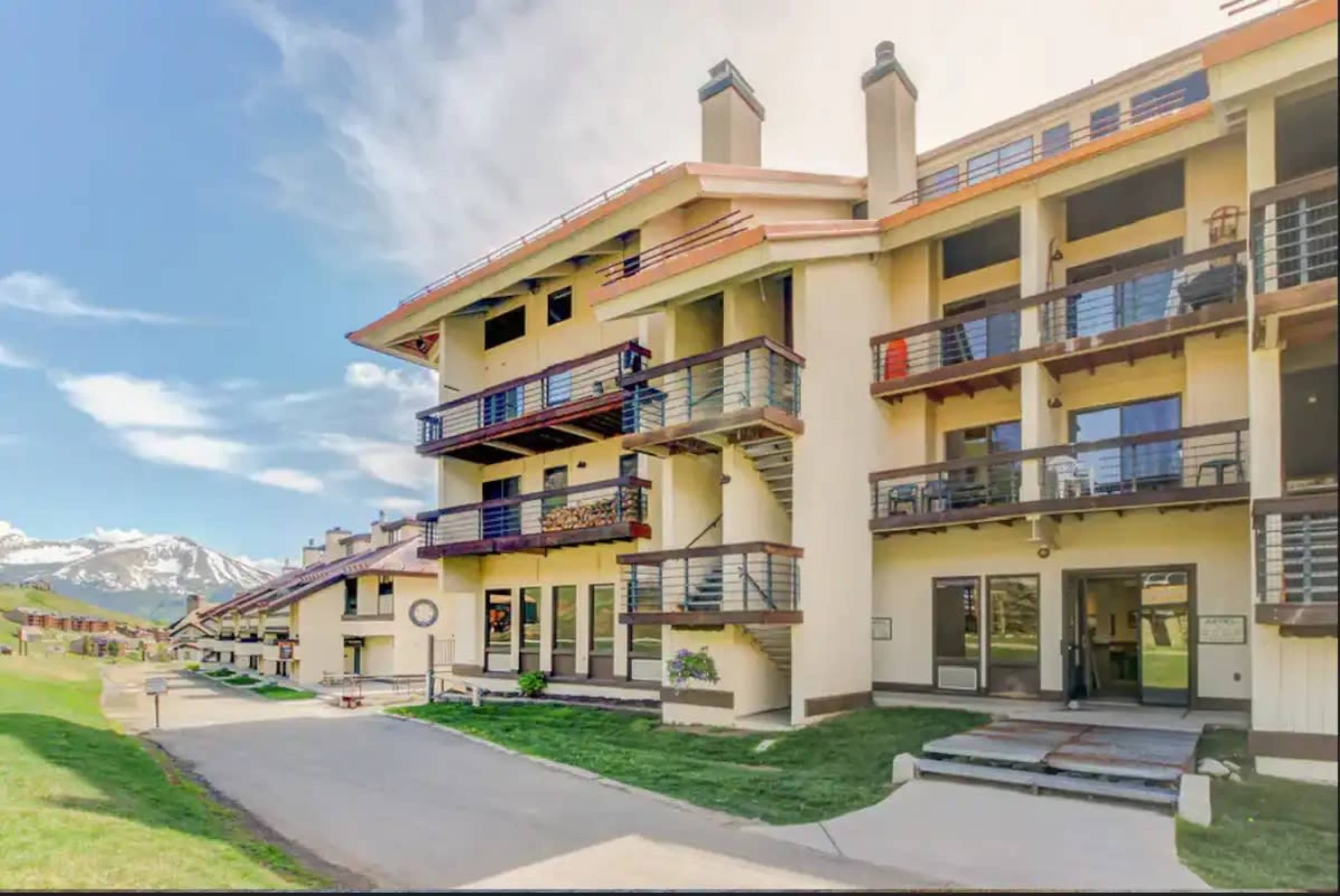 The image shows a row of multi-story buildings with balconies, set against a backdrop of mountains under a blue sky.