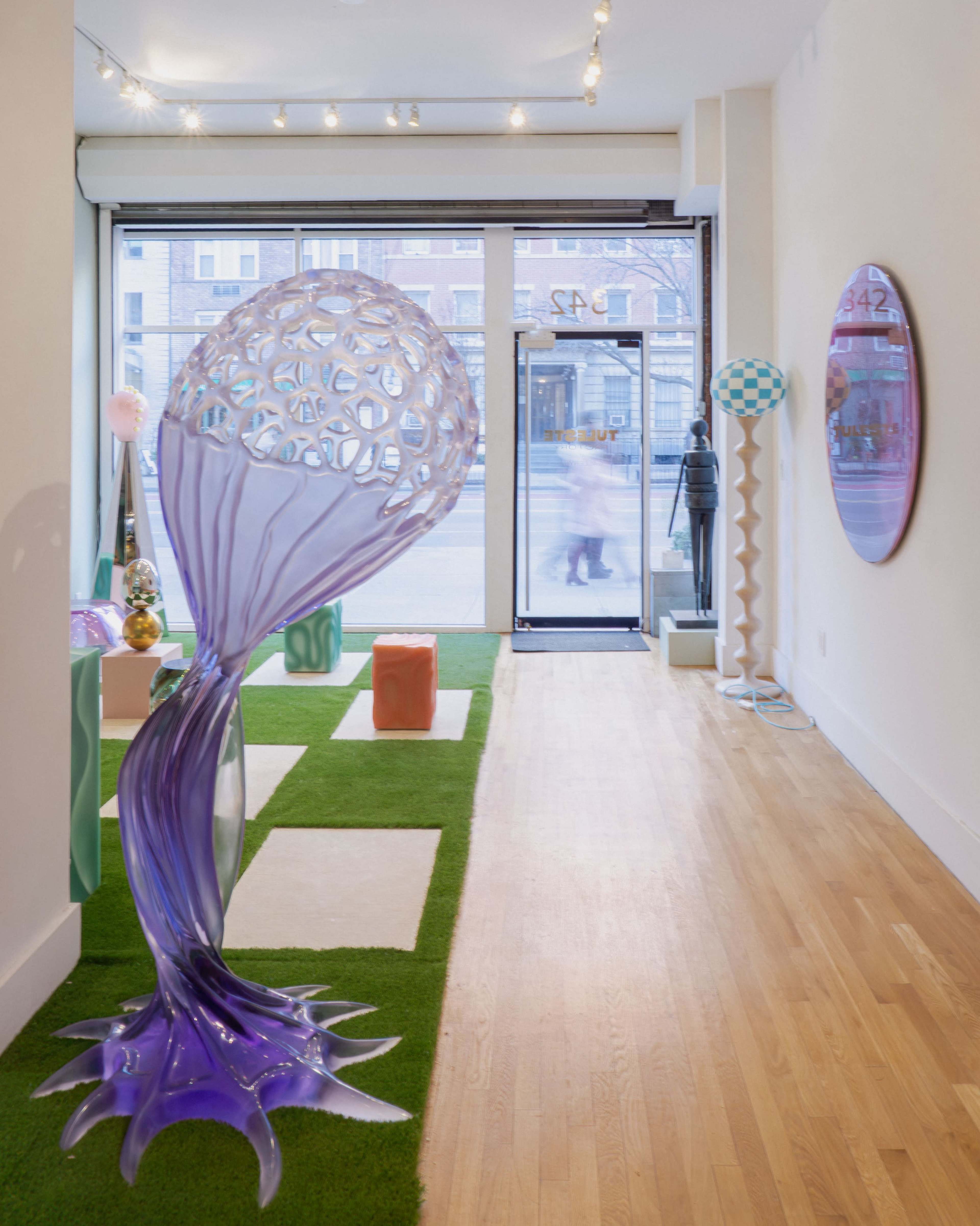 The image features an art gallery interior with a large, translucent sculpture resembling a tree in the foreground, and various colorful geometric shapes scattered on a green carpeted floor.