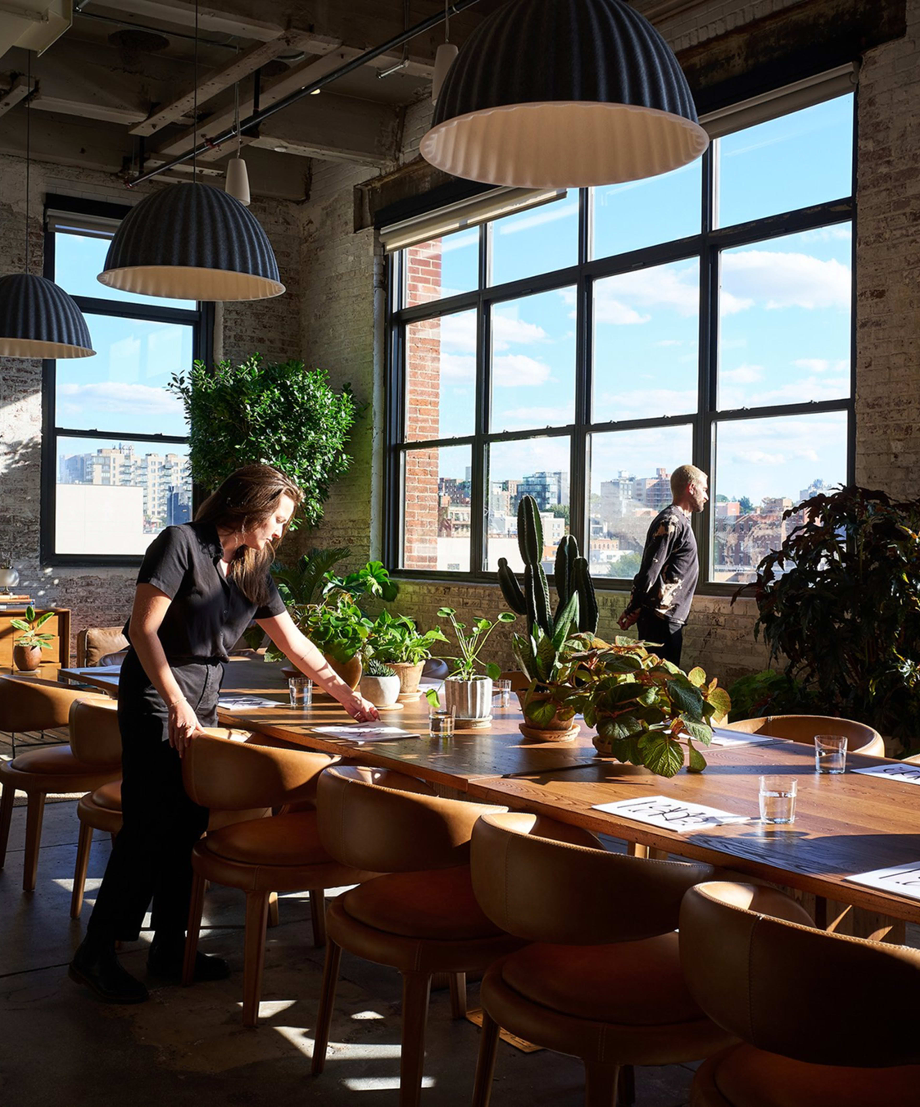 A person arranges plants on a long wooden table in a bright, industrial-style dining area with large windows.