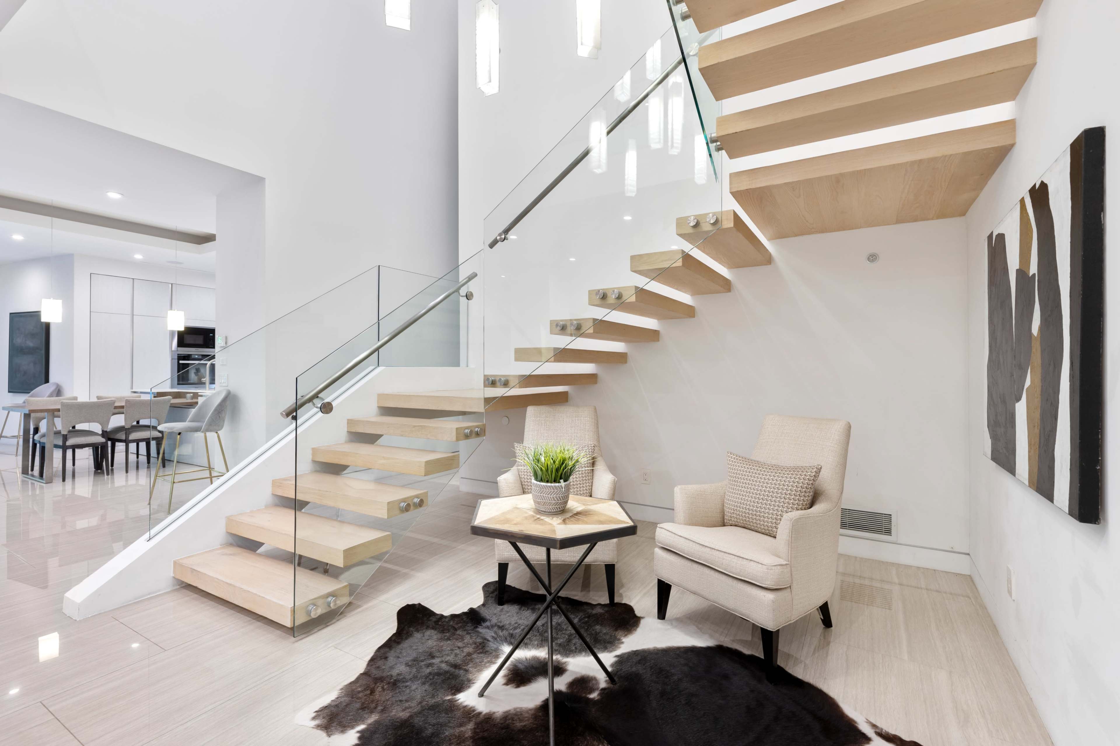 A modern staircase with wooden steps and glass railings leads to an upper level, alongside a cozy seating area with a patterned rug and a small plant on a side table.