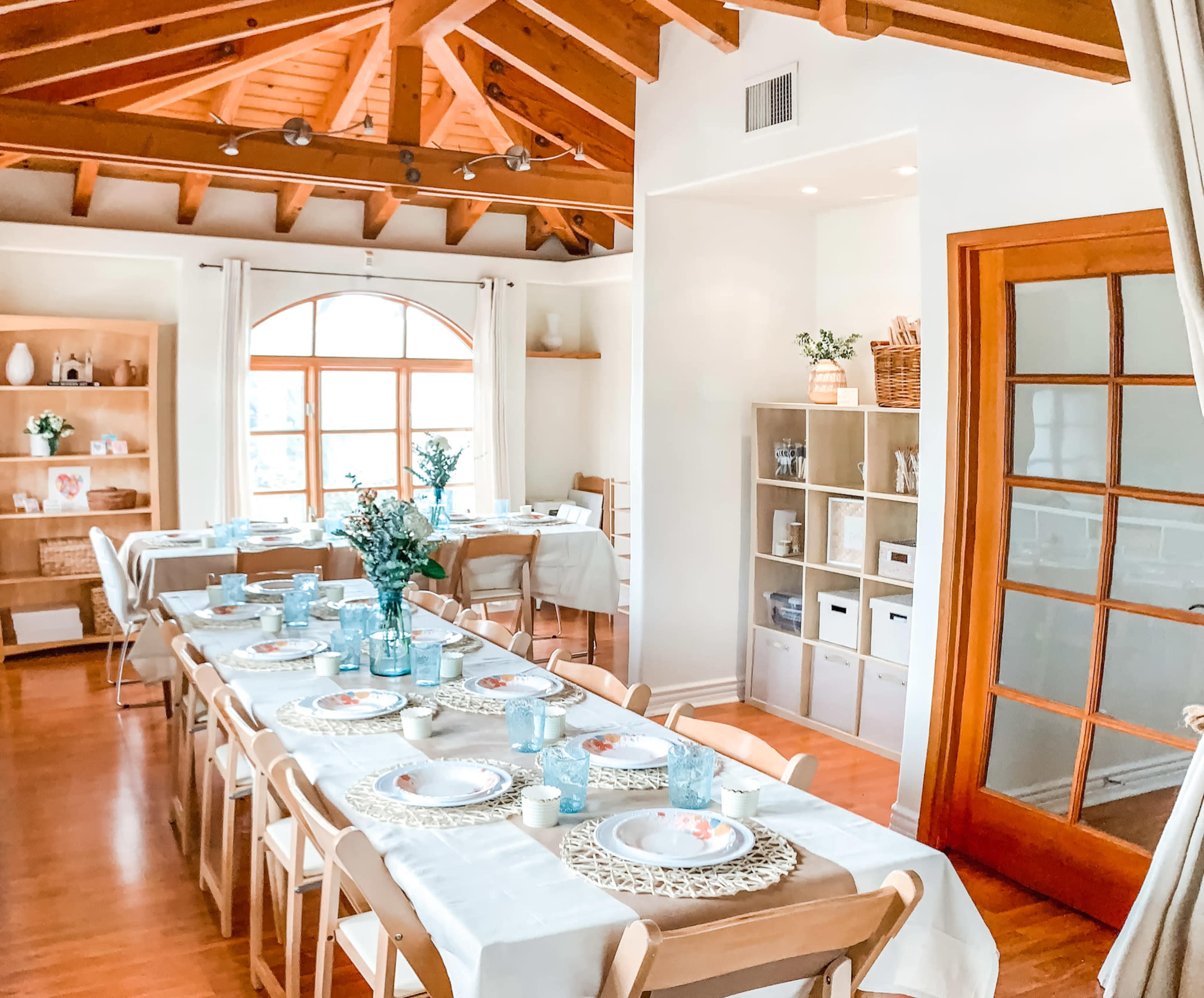 A bright dining area with wooden beams, a long table set with plates and glassware, and an adjacent shelving unit filled with decorative items.