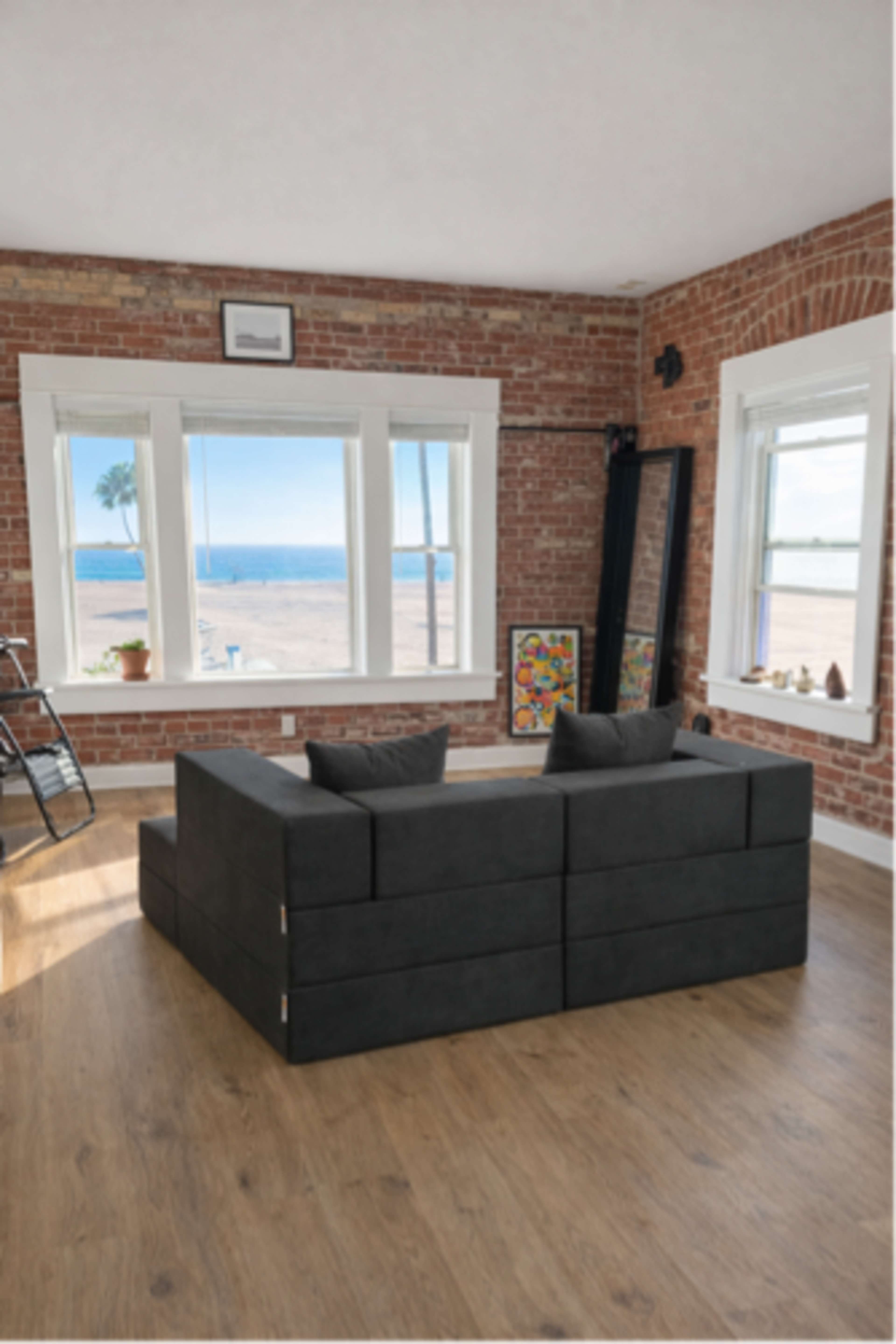 A black modular couch is positioned in a bright room with exposed brick walls, facing large windows that overlook a beach and ocean view.