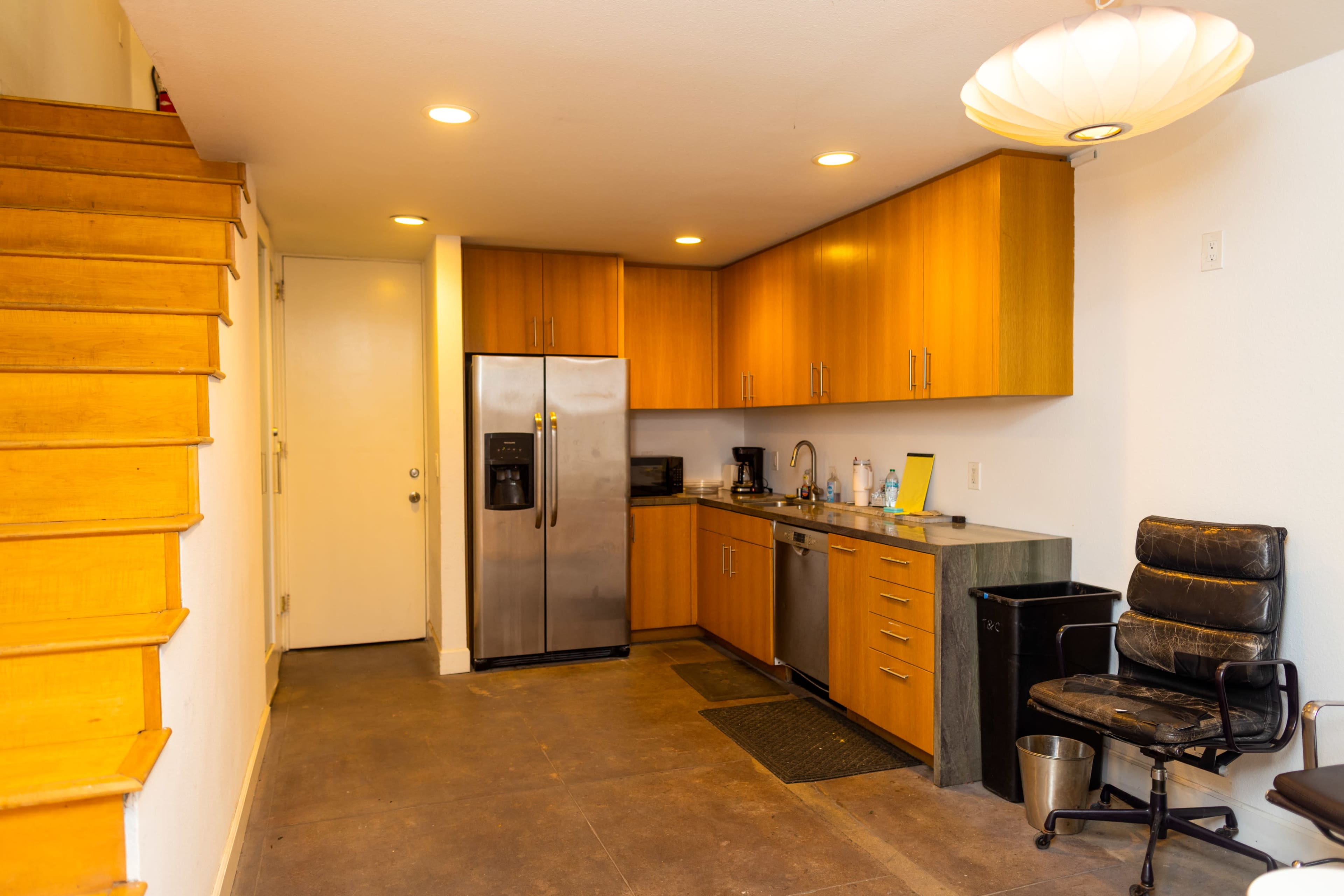The image shows a modern kitchen area with wooden cabinets, stainless steel appliances, and a staircase leading to an upper level.