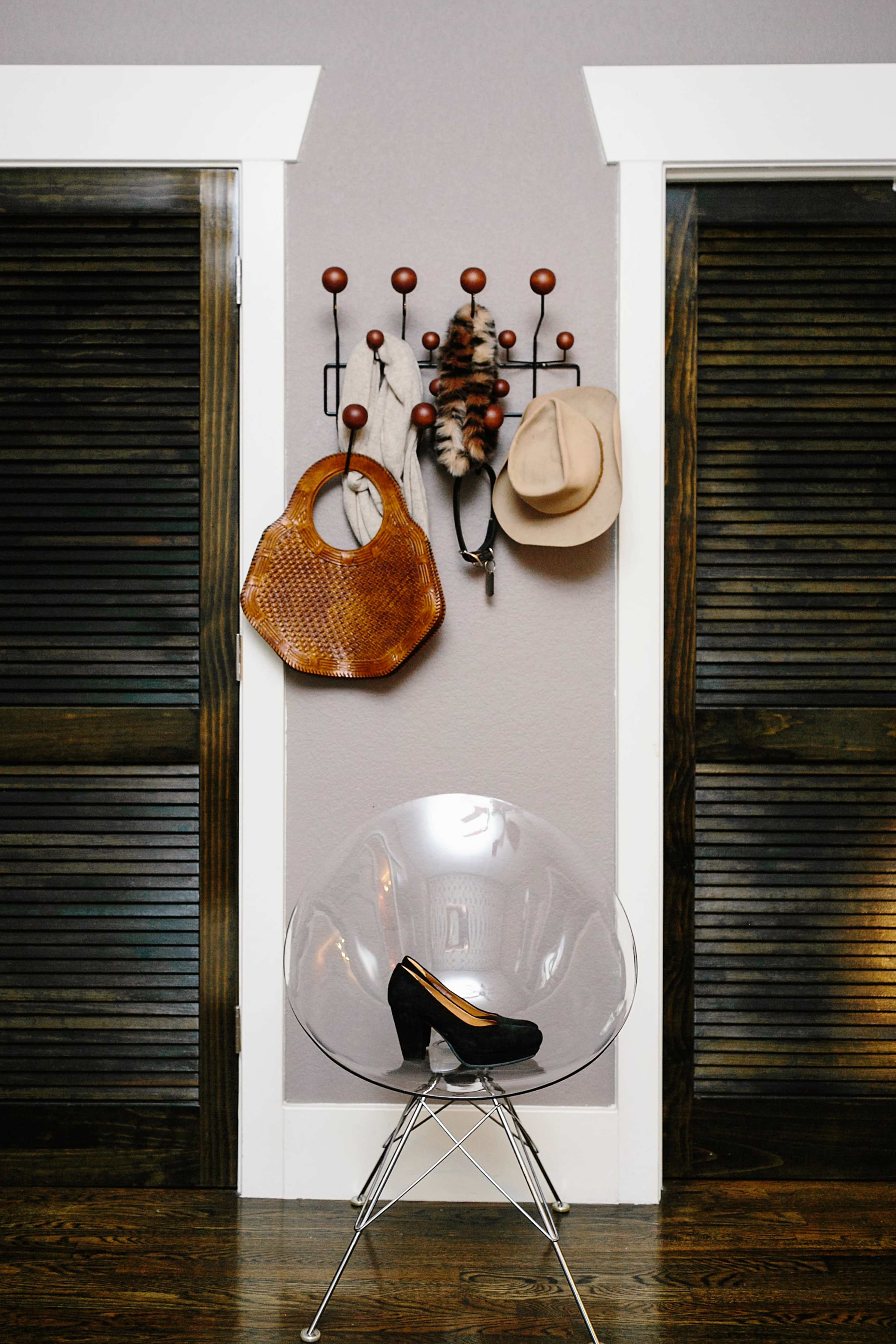 A black high-heeled shoe rests on a transparent chair in front of two closed wooden doors, with accessories hanging on a rack on the wall.