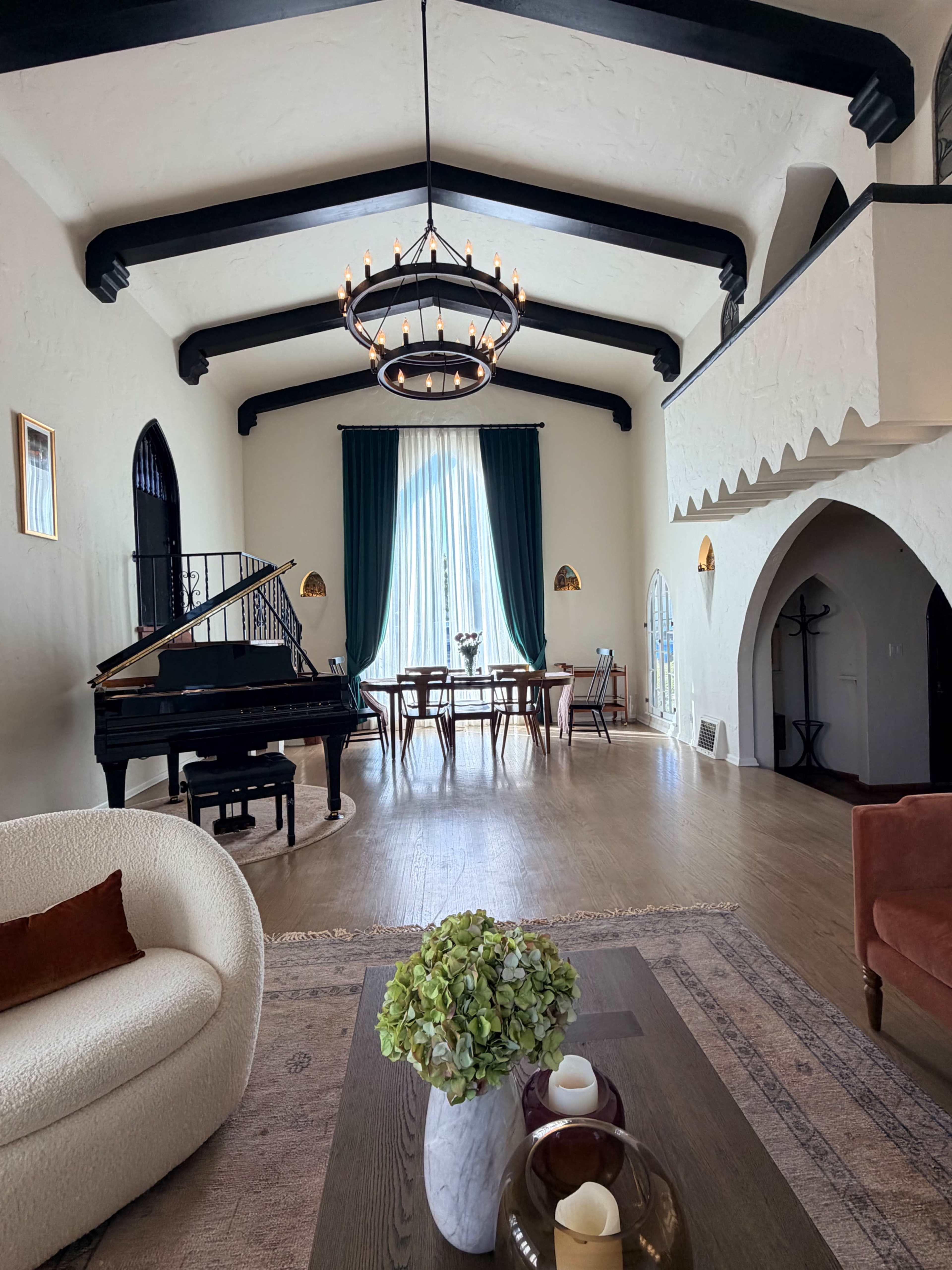 The image depicts a spacious interior of a home with high ceilings, featuring a grand piano, a dining area with a table, and large windows adorned with drapes.