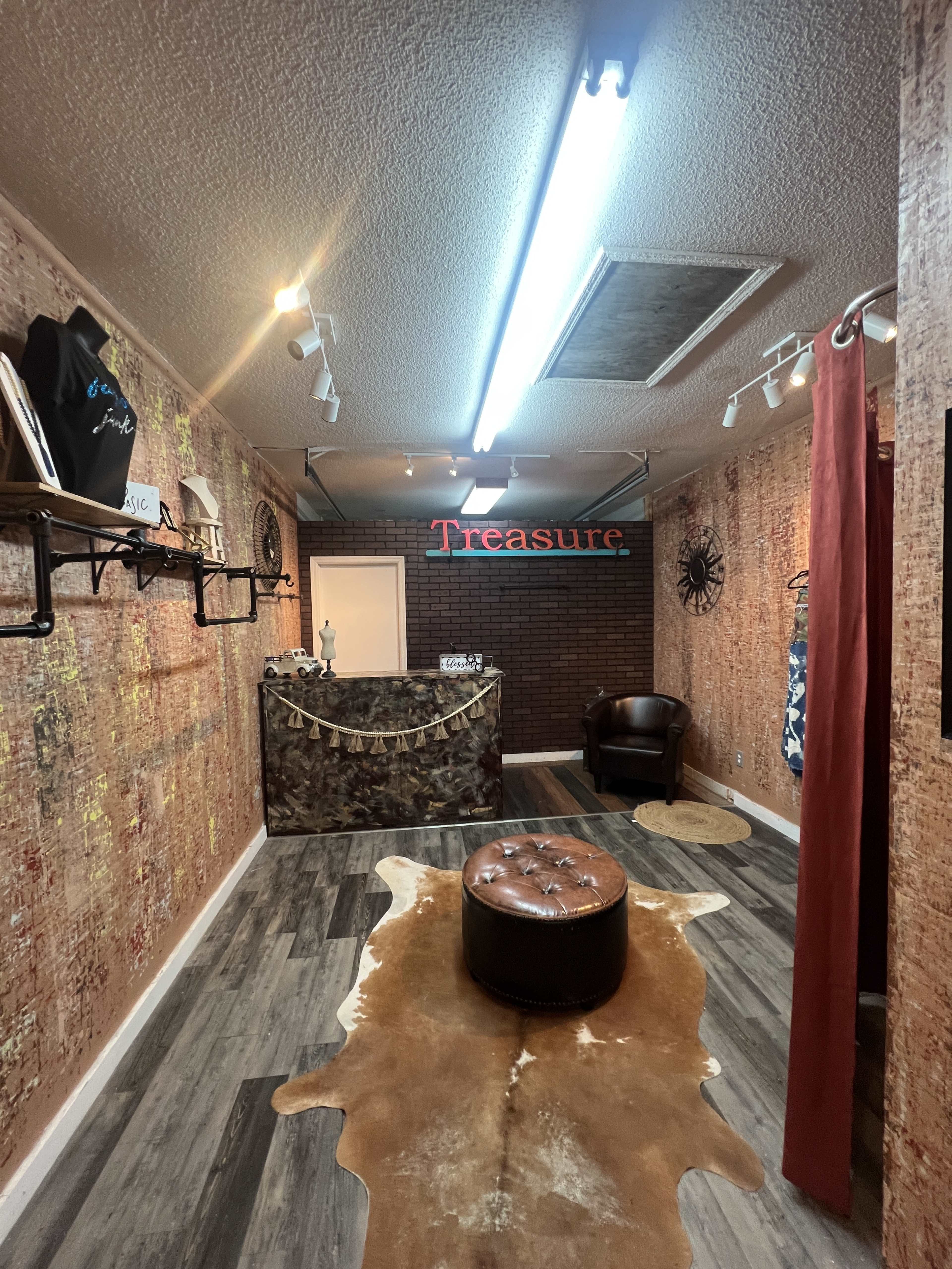 The image depicts an interior of a small retail space featuring a decorative wall, a reception desk with the word "Treasure," a brown leather chair, and a circular area rug.