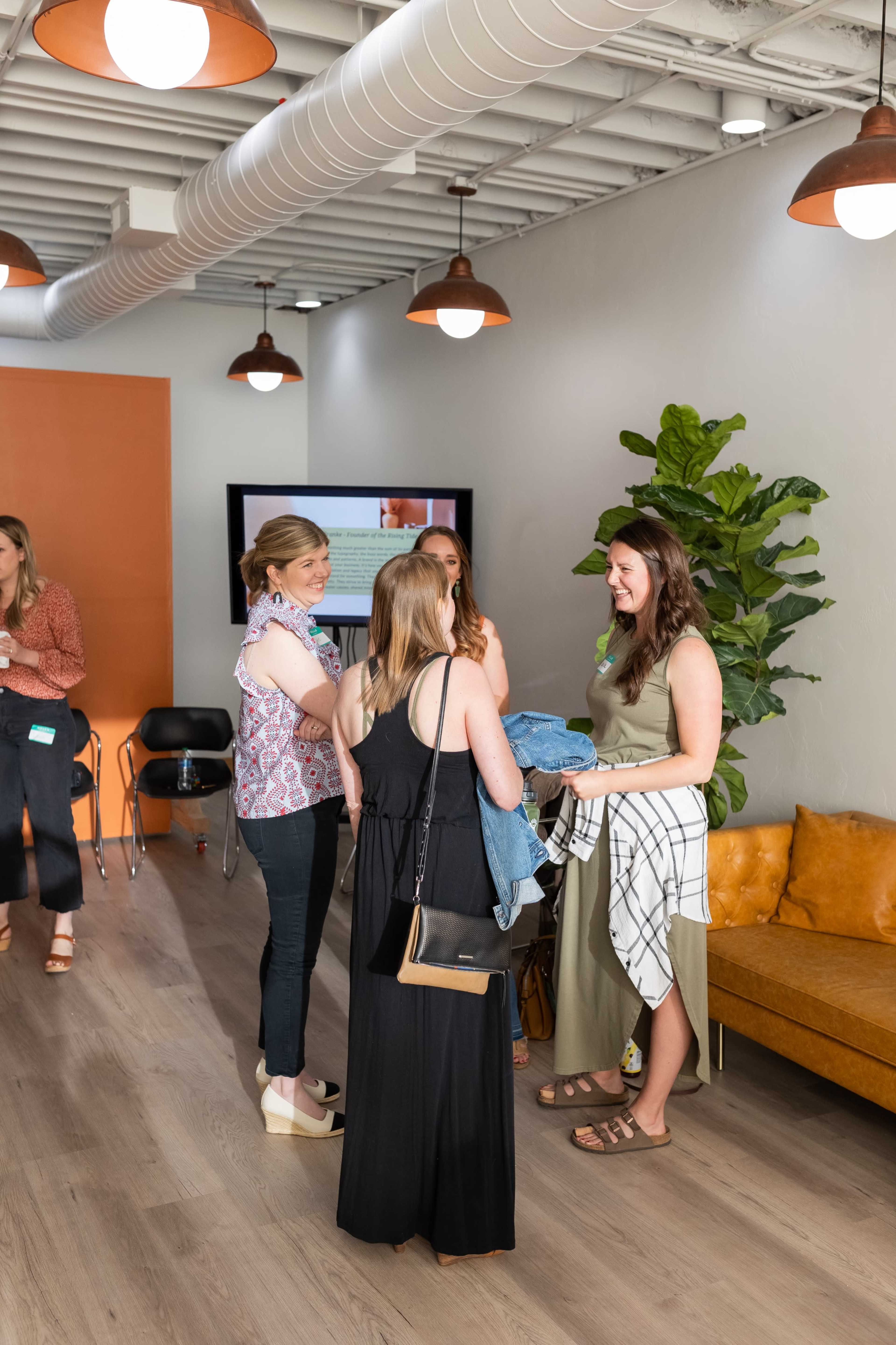 A group of five women chat and interact in a modern, well-lit lounge area with plants and a large screen in the background.
