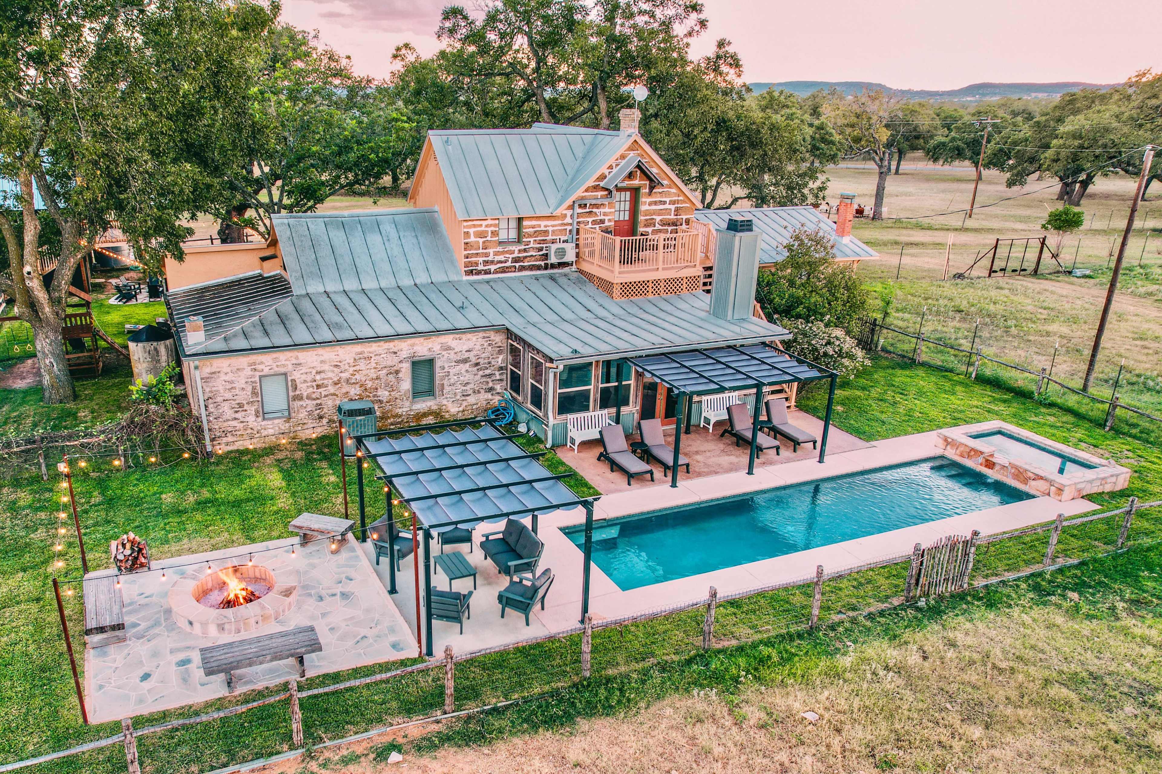 The image shows a modern house with a stone exterior, a swimming pool, a fire pit area, and a fenced backyard surrounded by trees.