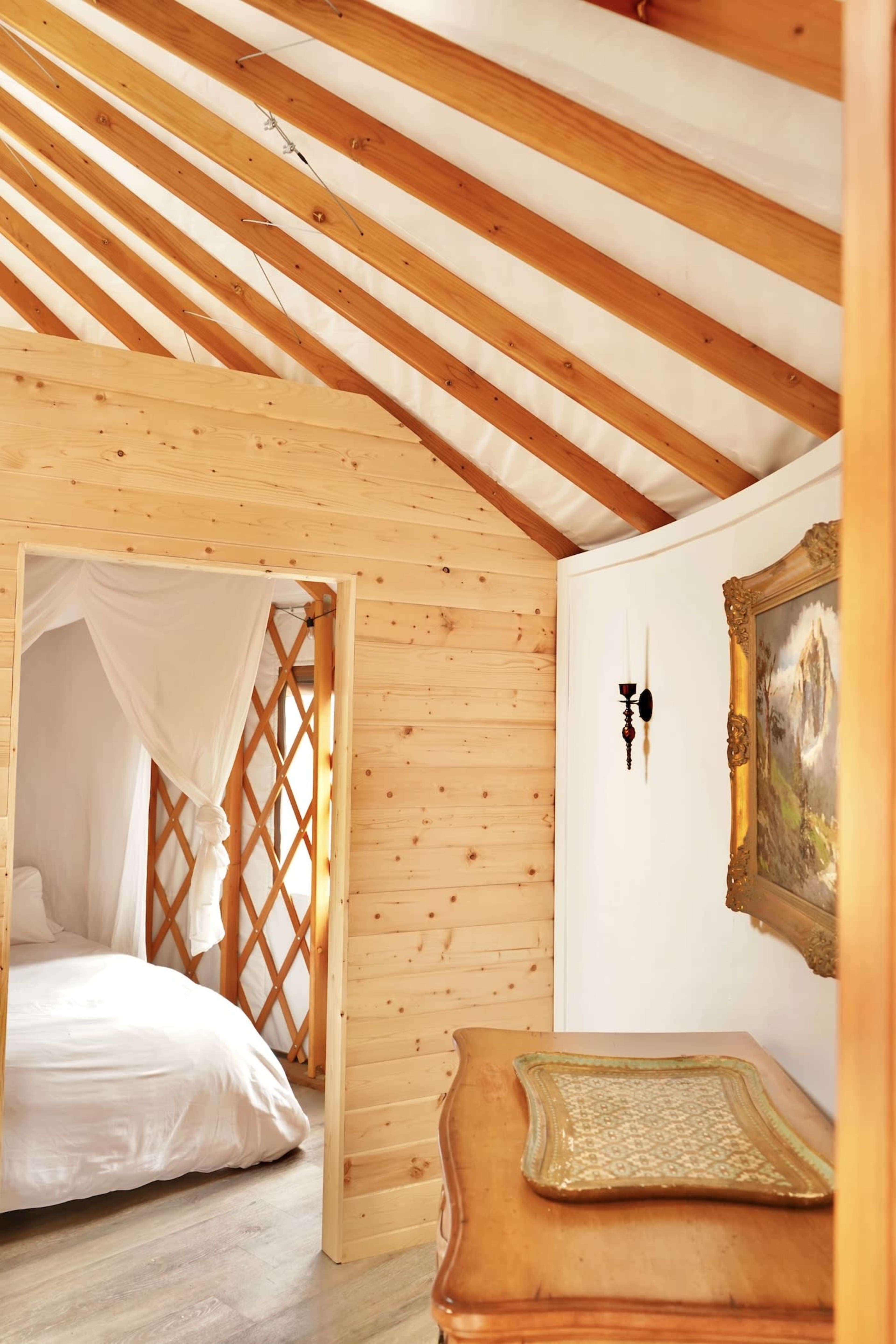 A cozy interior of a wooden room with a structured ceiling, featuring a small bed, a vintage painting, and a wooden table.