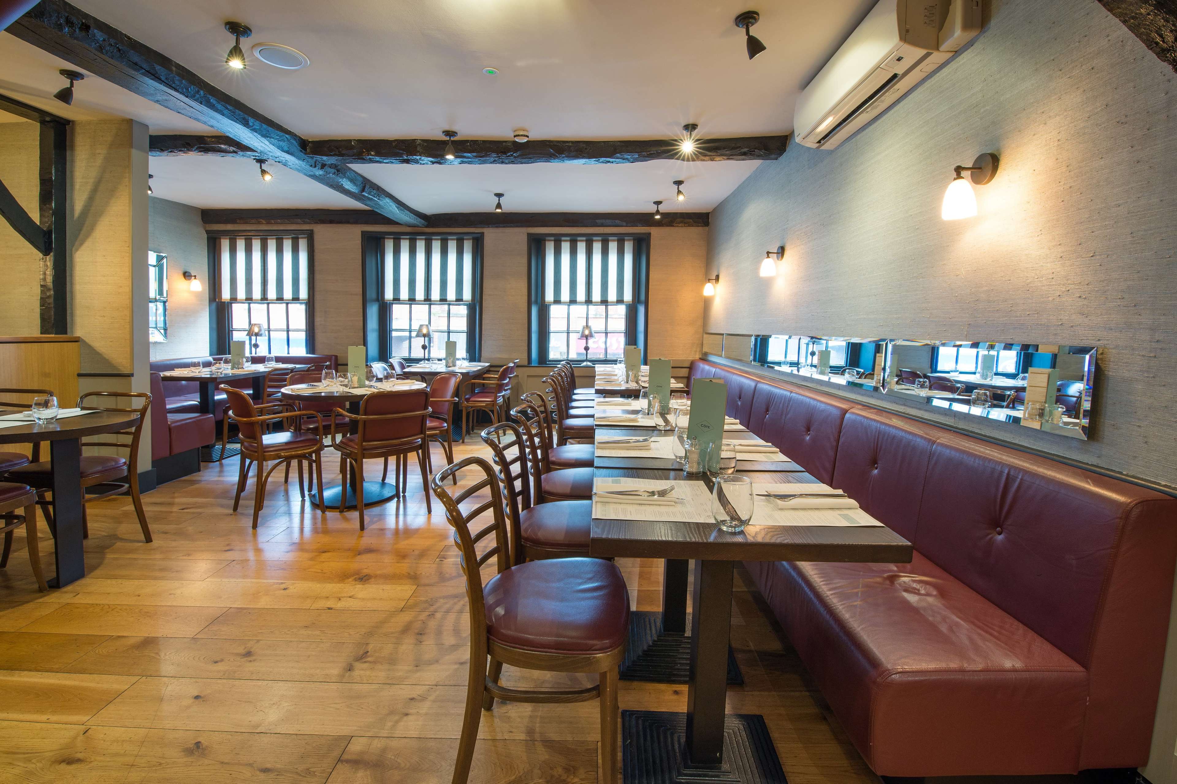 The image shows an interior view of a restaurant featuring wooden floors, red upholstered seating, and tables arranged for dining under soft lighting.