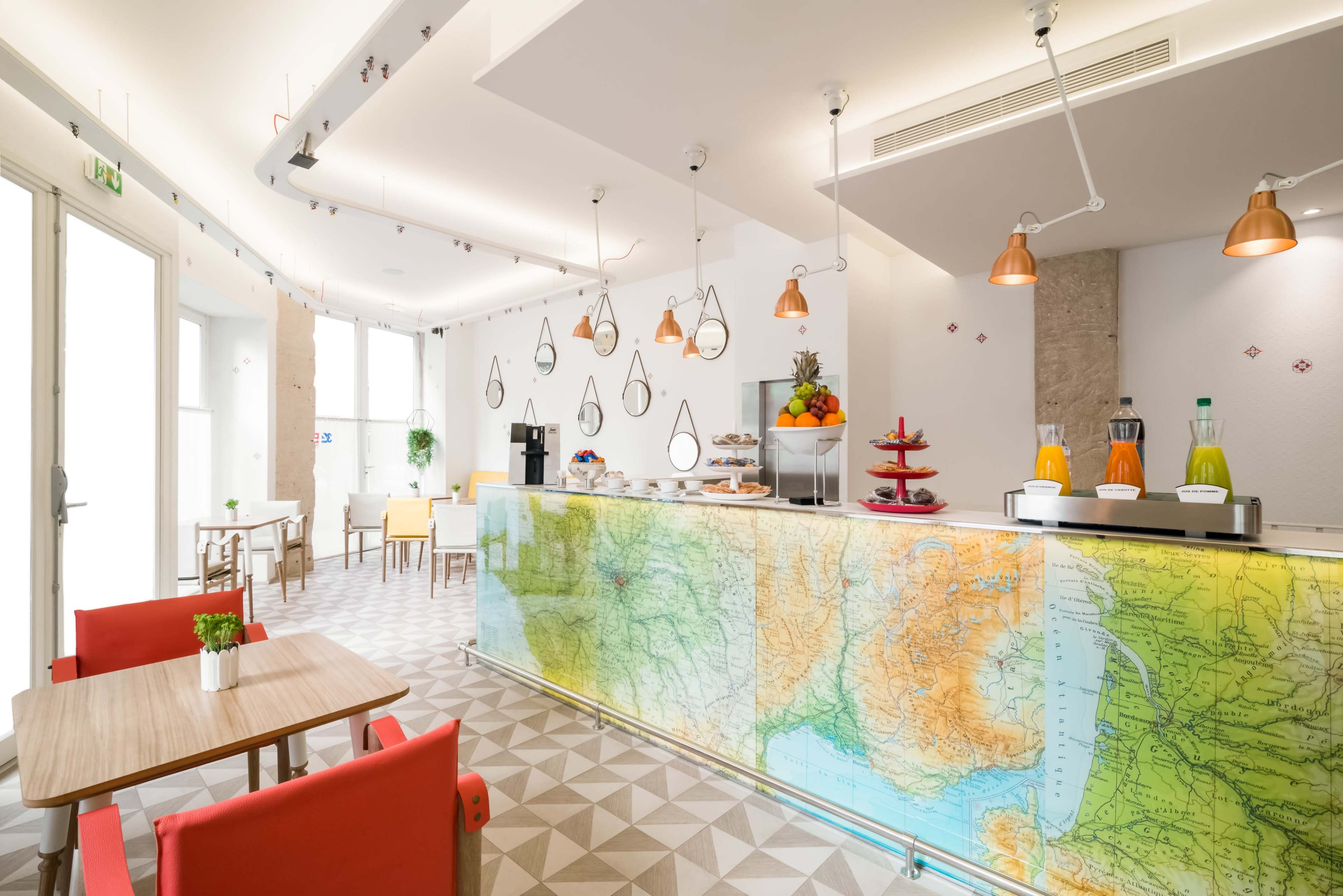 A café interior features a map-themed countertop, wooden tables, and colorful pendant lights, with a display of beverages and pastries.