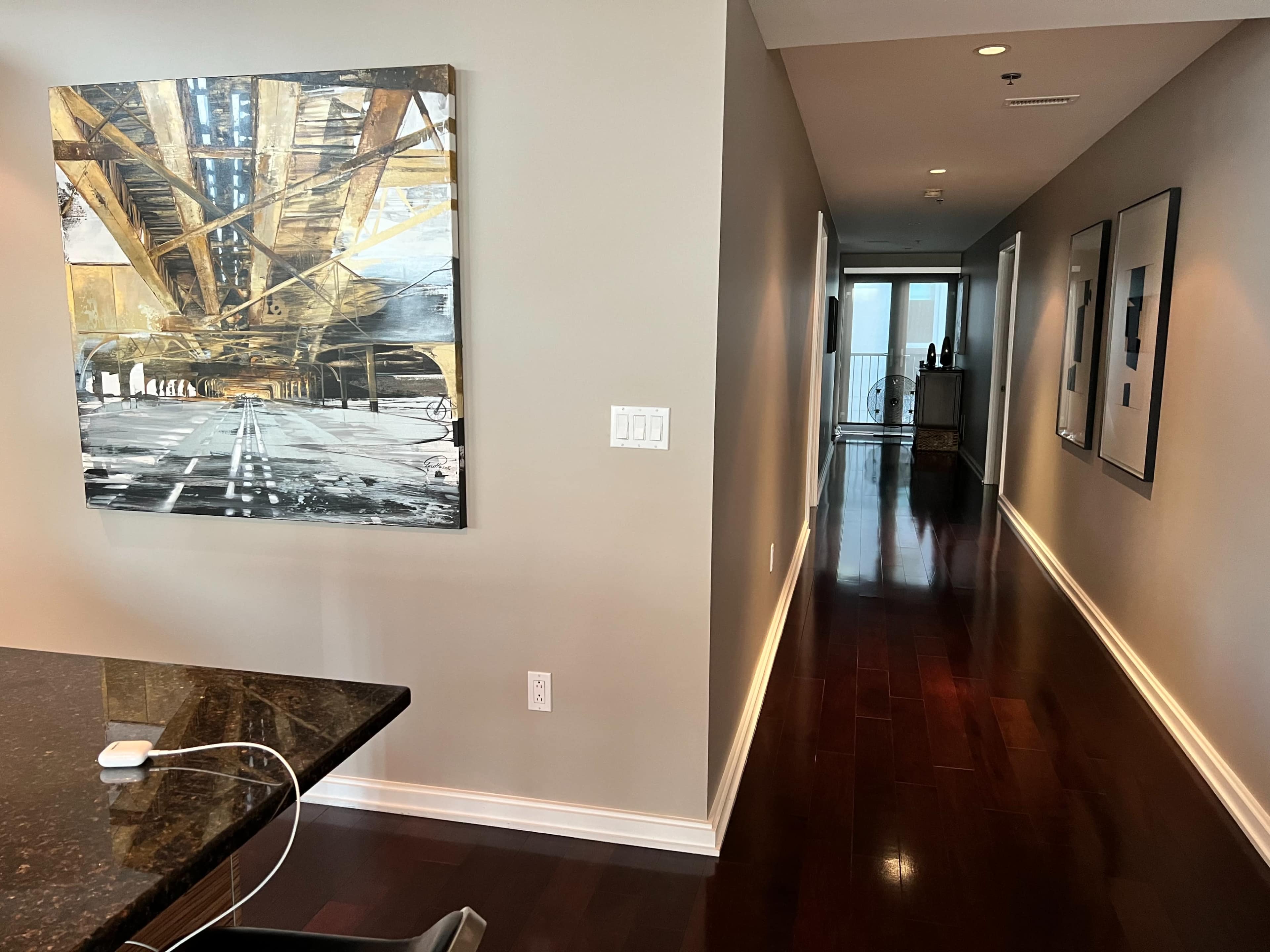 A modern interior space featuring a hallway with glossy wooden floors and a large painting on the wall above a counter, leading to a glass door at the end.