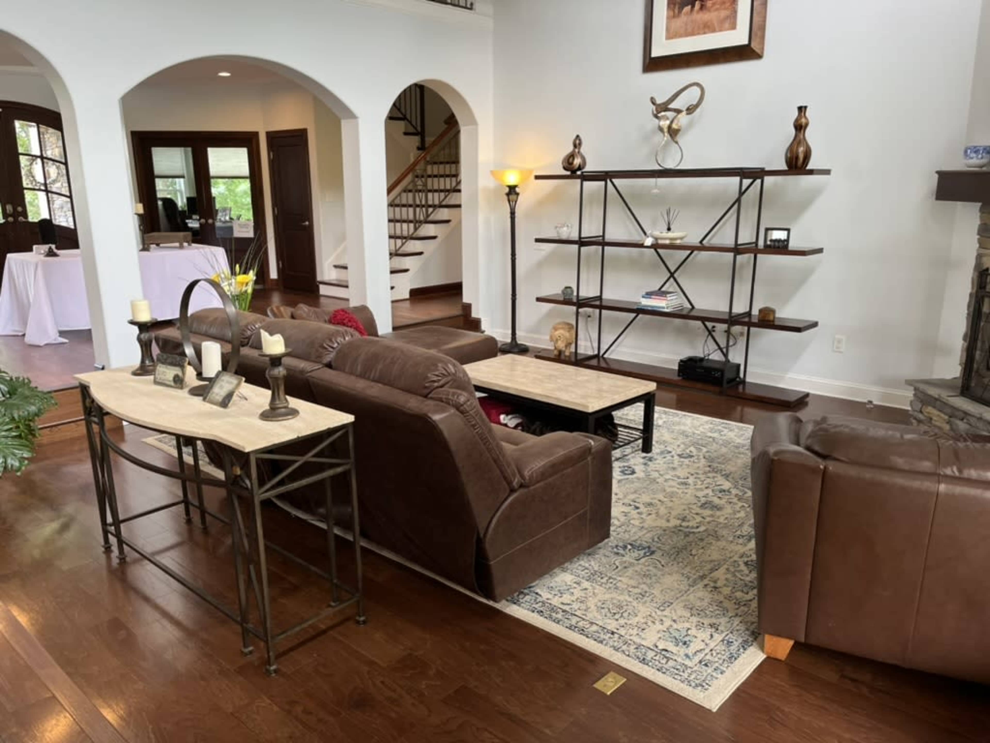 The image shows a spacious living room with brown couches facing a decorative shelf and a fireplace, featuring wooden flooring and large windows.