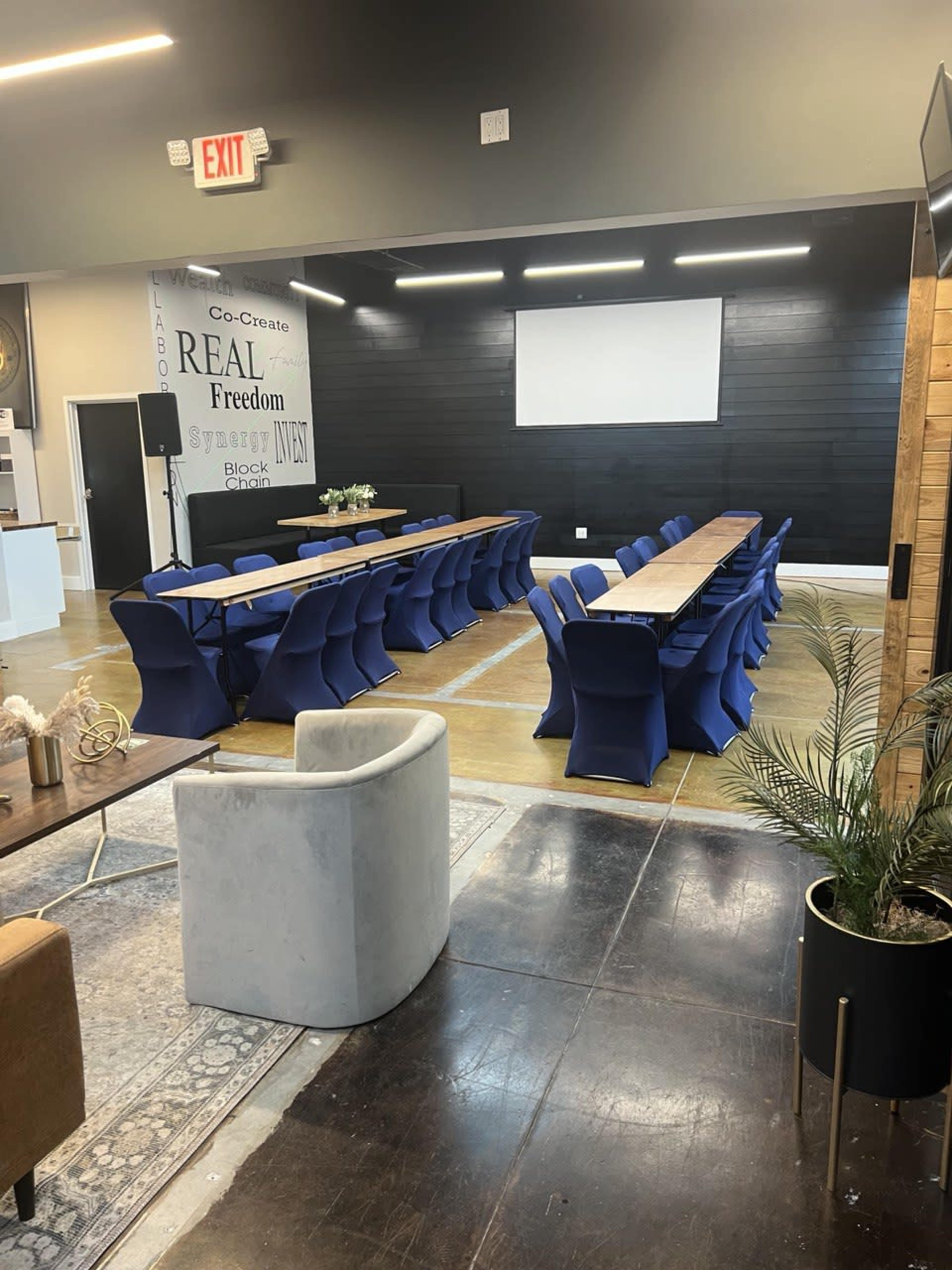 A conference room setup features a long table surrounded by blue chairs, with a screen on the wall and decor elements like plants and a rug nearby.