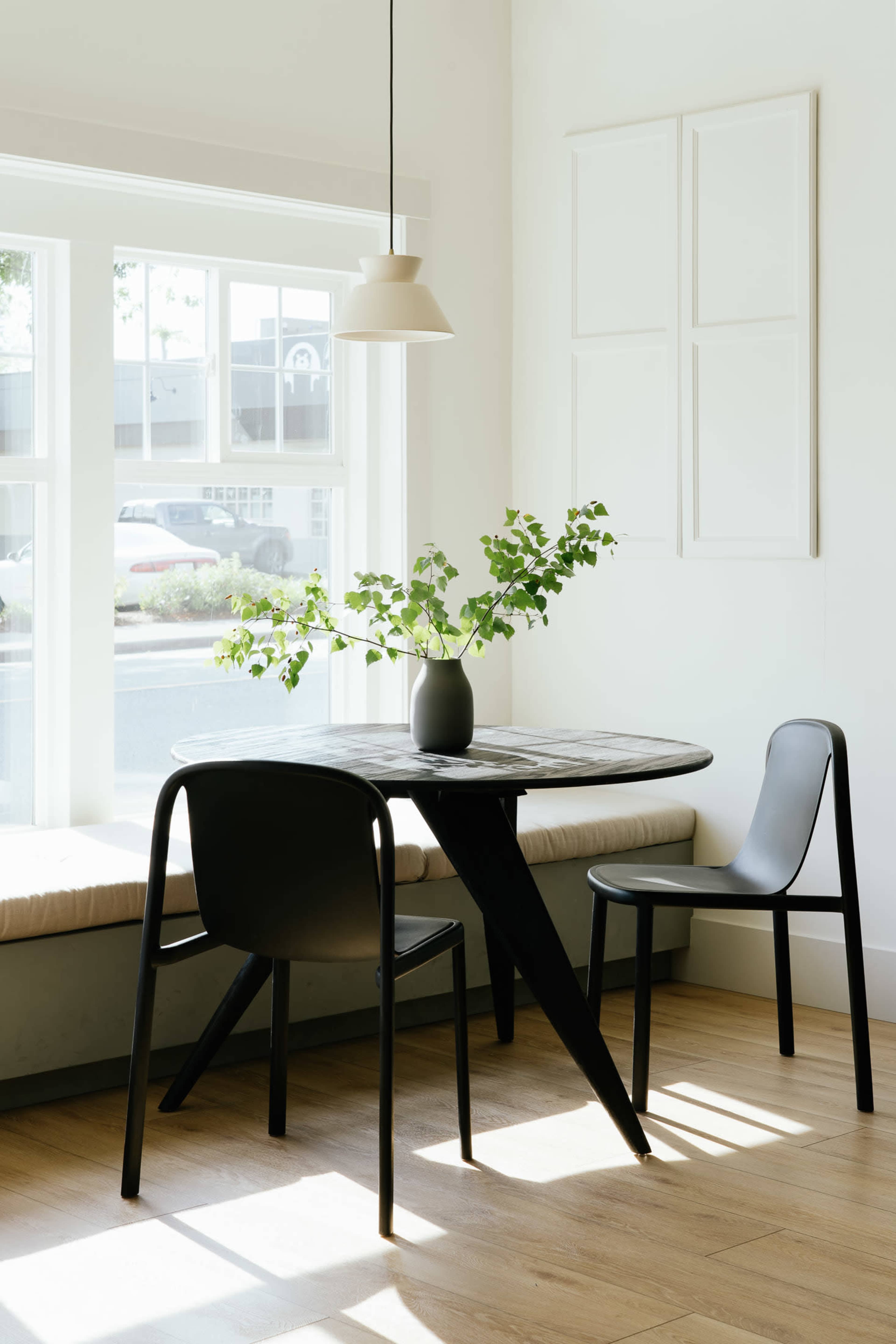 A small table with a vase of greenery is positioned near a window in a bright, minimalist room.