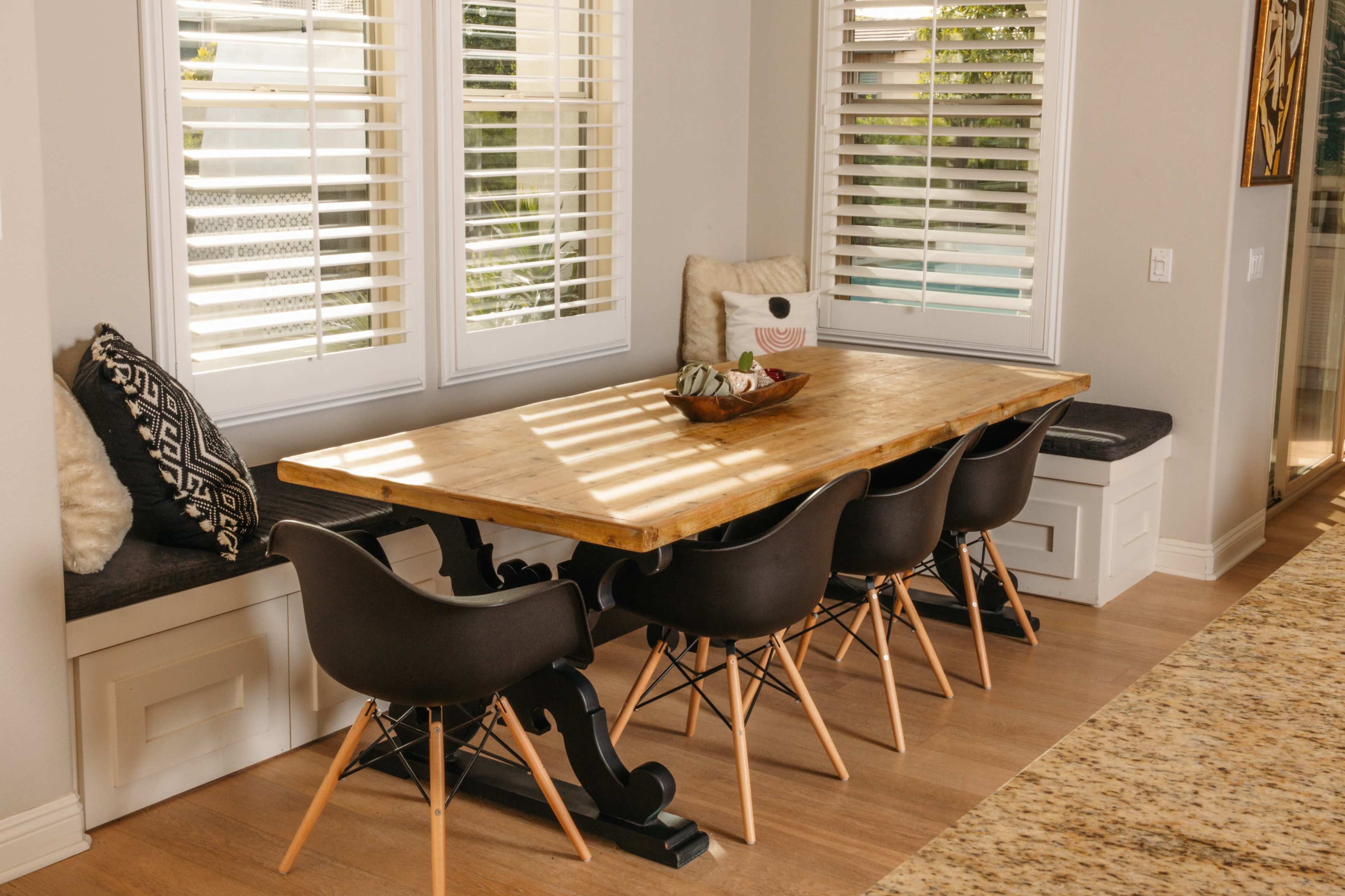 A wooden dining table with black chairs is positioned near windows with white shutters in a bright room.