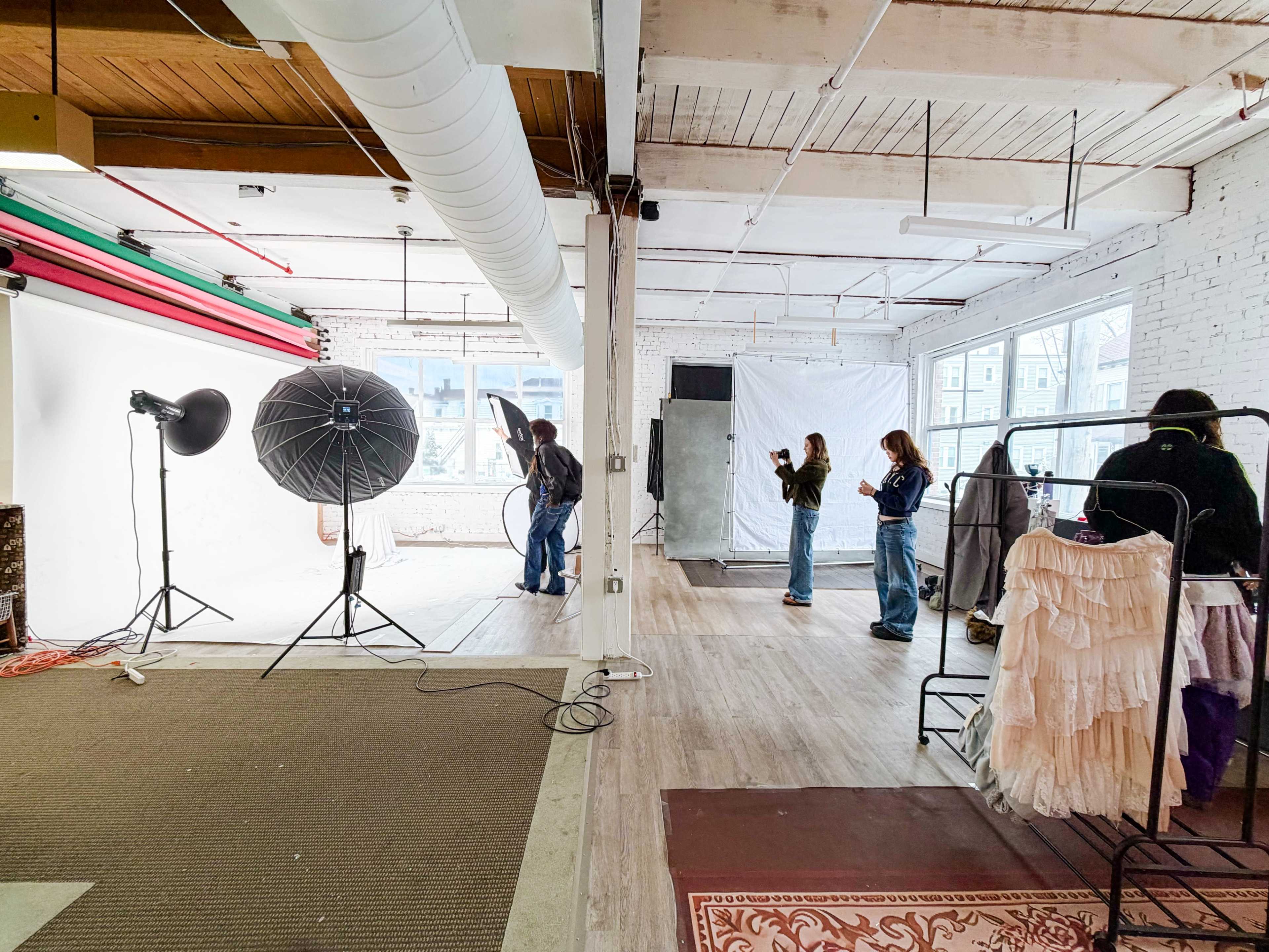 A photography studio features several people working with lights and backdrops while styling garments on clothing racks.