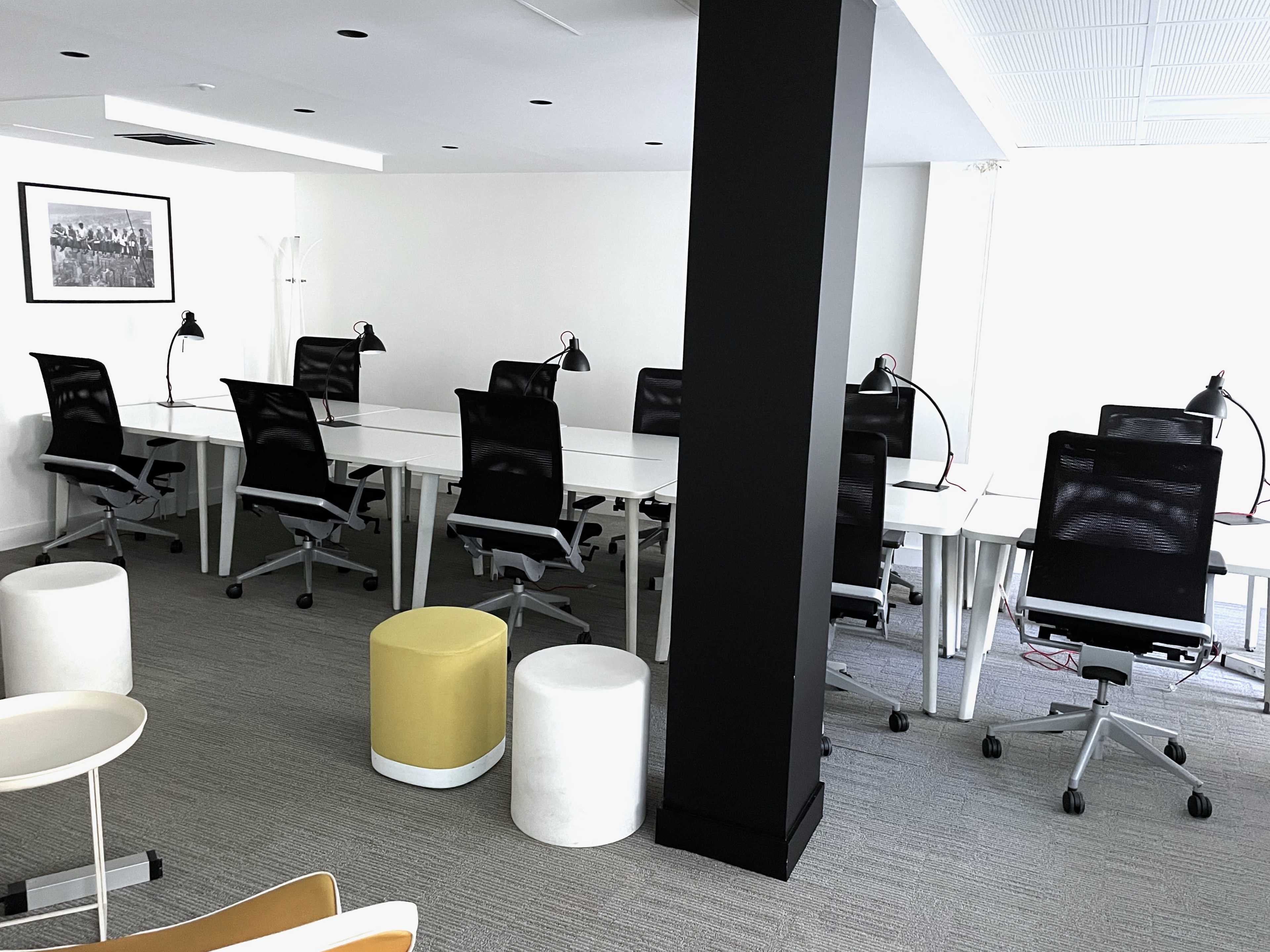 A modern office space featuring a row of sleek desks with black office chairs, accompanied by several standing lamps and a few round stools.