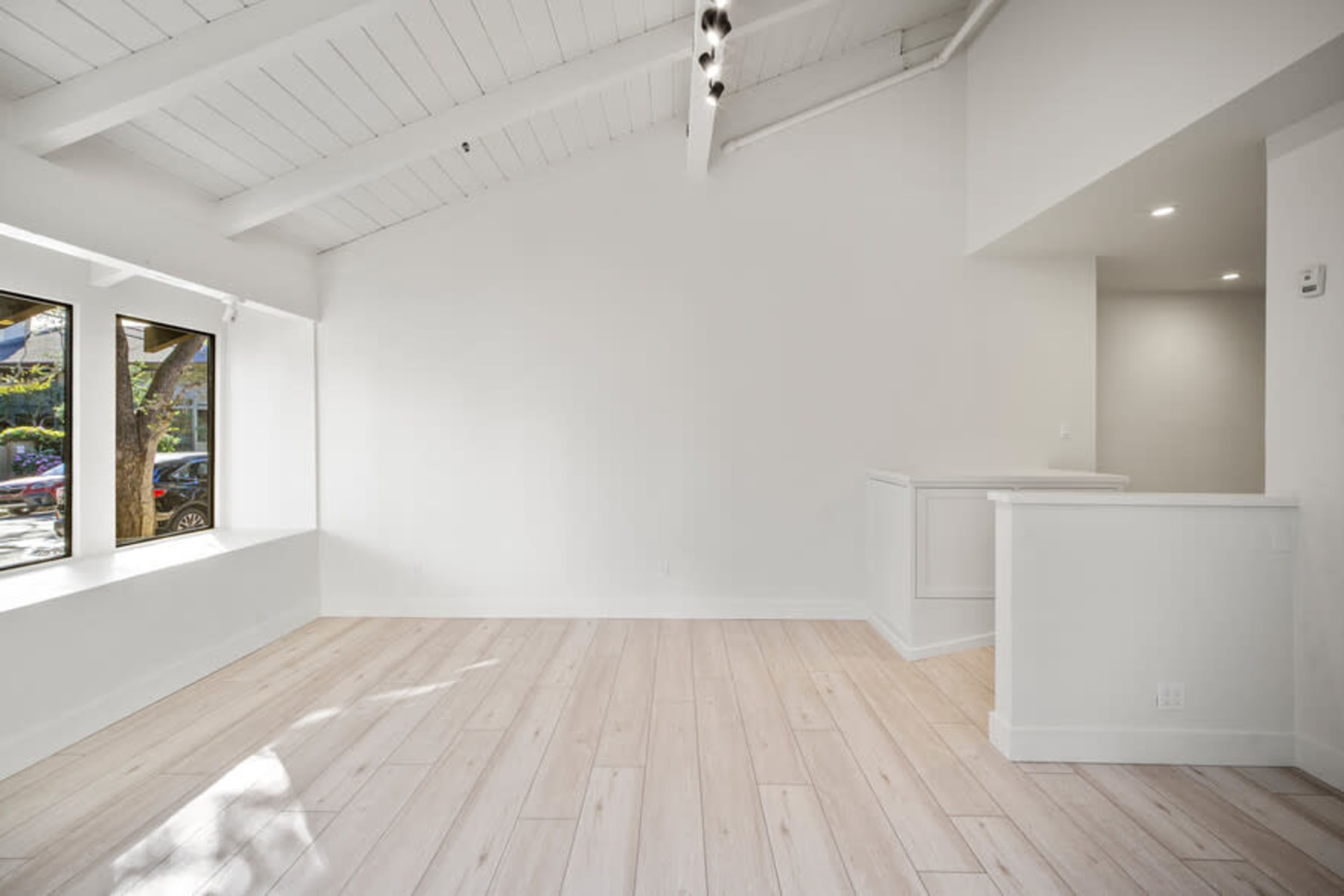 A bright, empty room features high, white-beamed ceilings, large windows, and light-colored flooring.