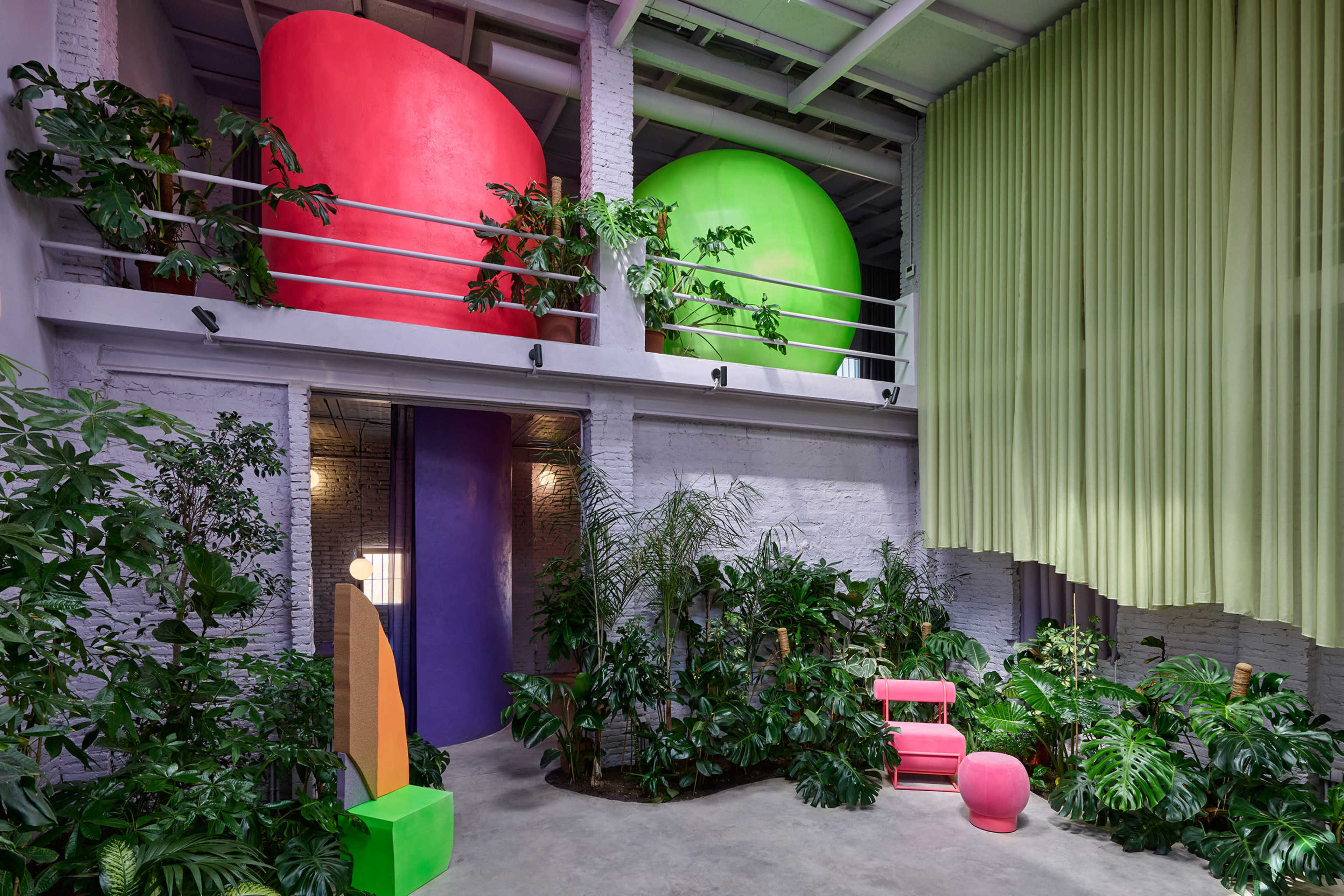 The image shows a brightly colored interior space featuring large, rounded forms in pink and green, surrounded by various indoor plants and complemented by a purple doorway and green drapery.