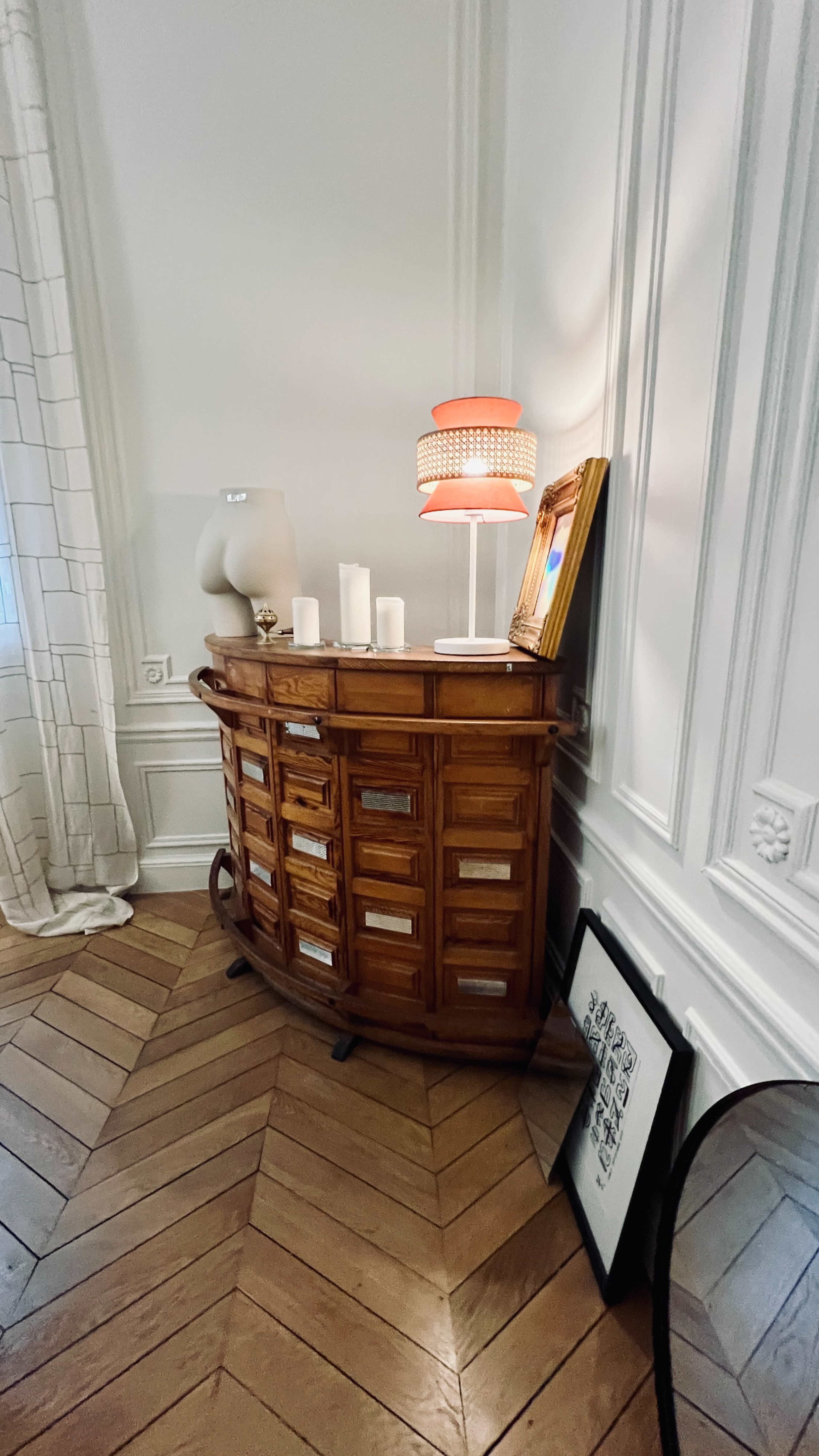 A wooden cabinet with multiple drawers is positioned in a corner of a room, accompanied by a lamp, candles, and framed artwork on a wooden floor.