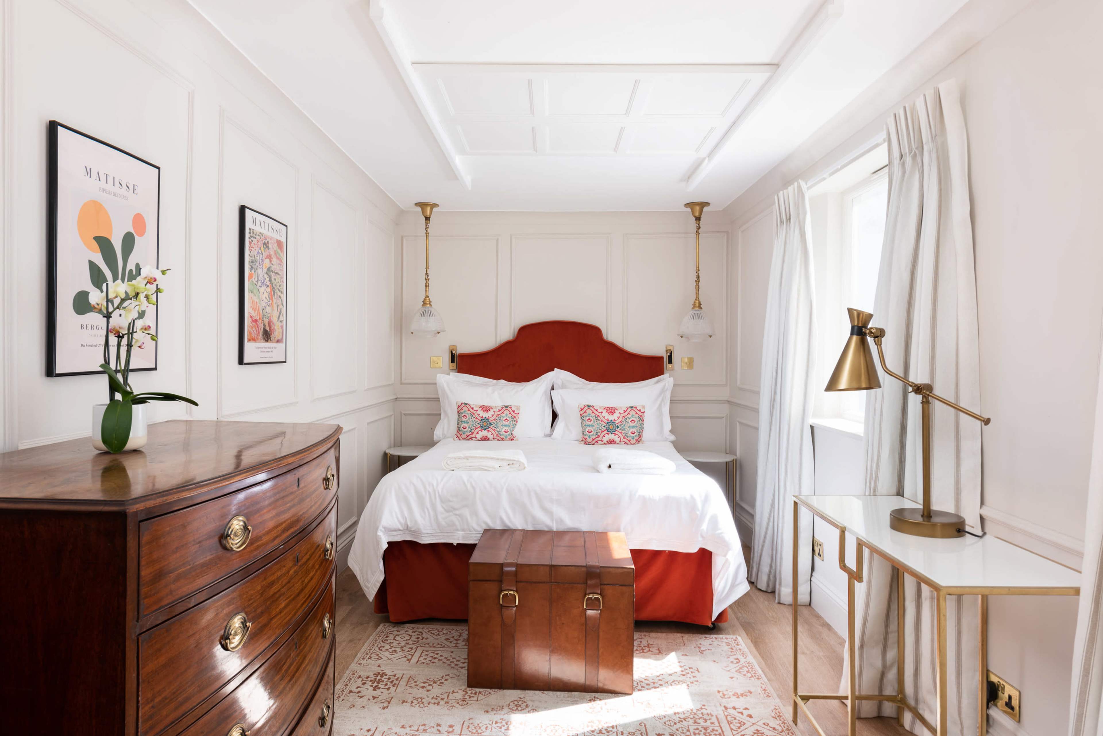 A cozy bedroom featuring a red-tufted headboard, white bedding, a wooden dresser, and a decorative chest at the foot of the bed.