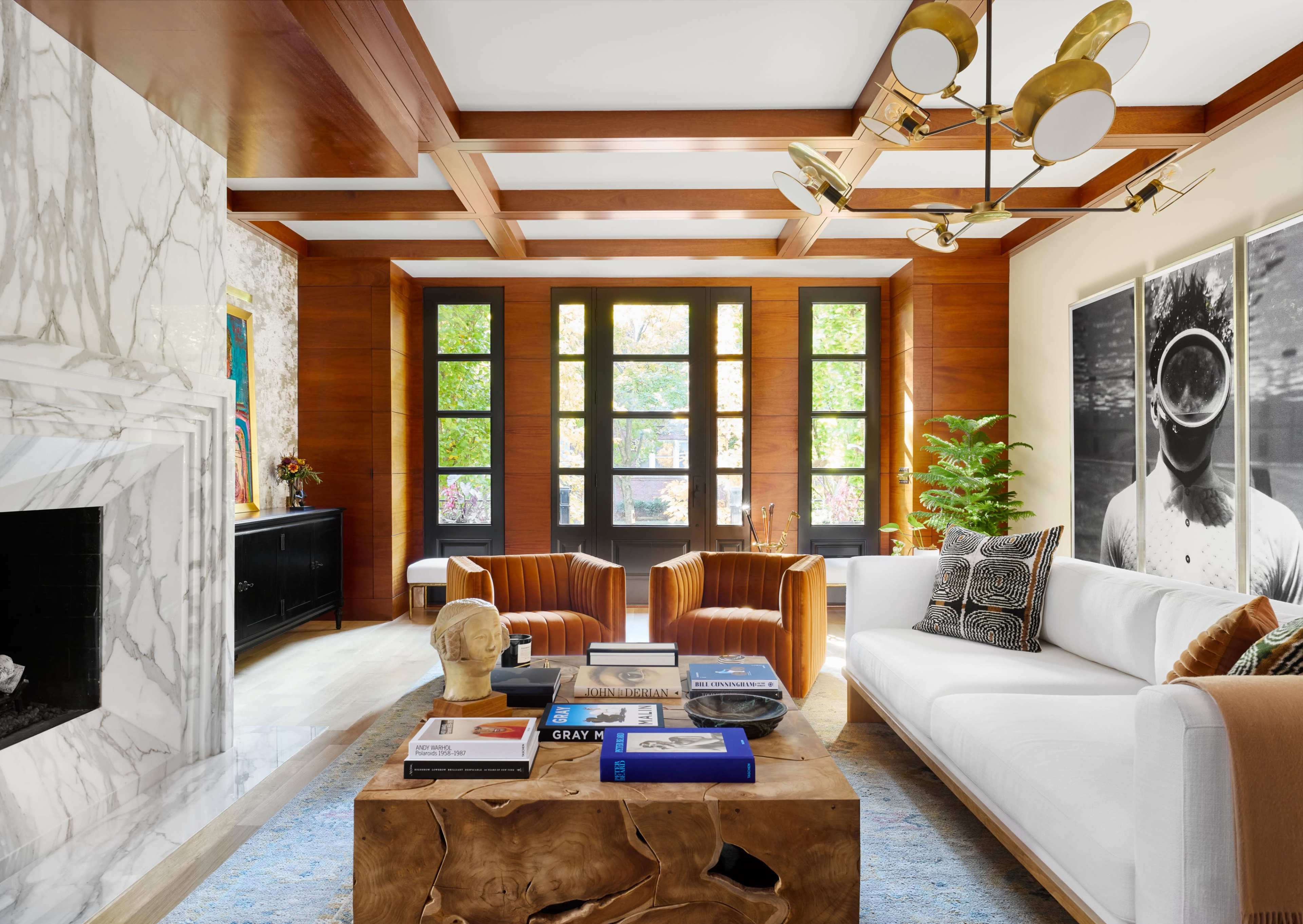 The room features a white sofa, two brown leather chairs, a wooden coffee table with various books, and large windows overlooking a garden.