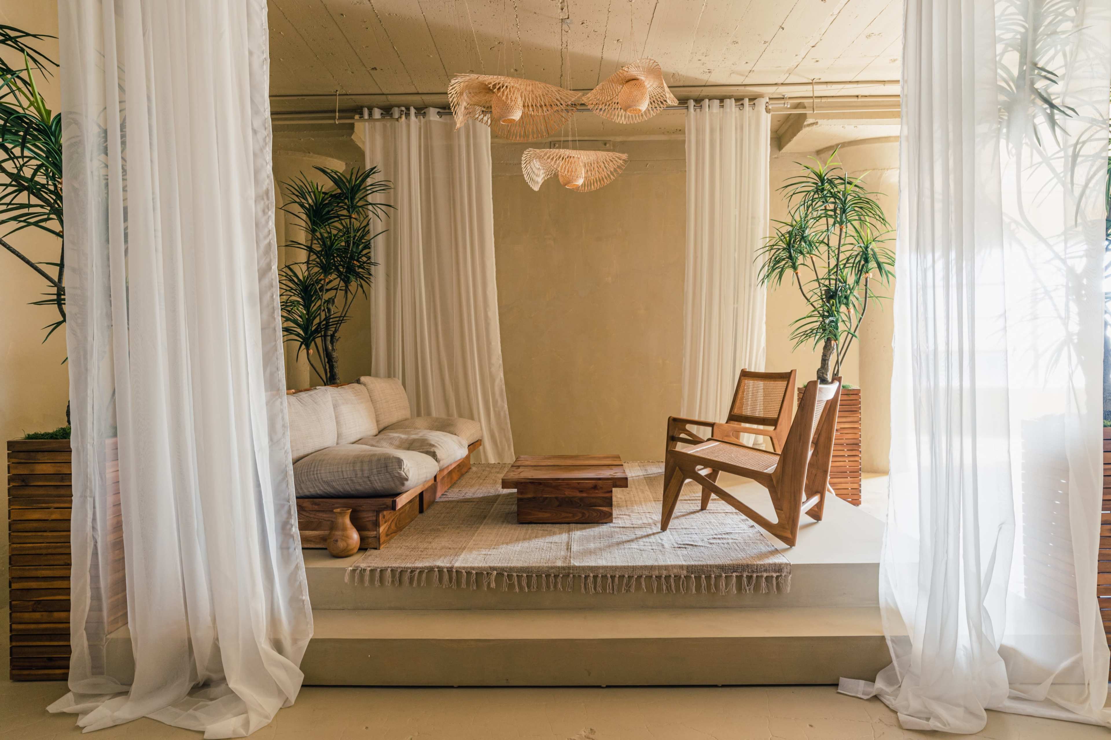 The image depicts a cozy, minimalistic living space with a wooden sofa, a coffee table, and large potted plants, surrounded by sheer white curtains.