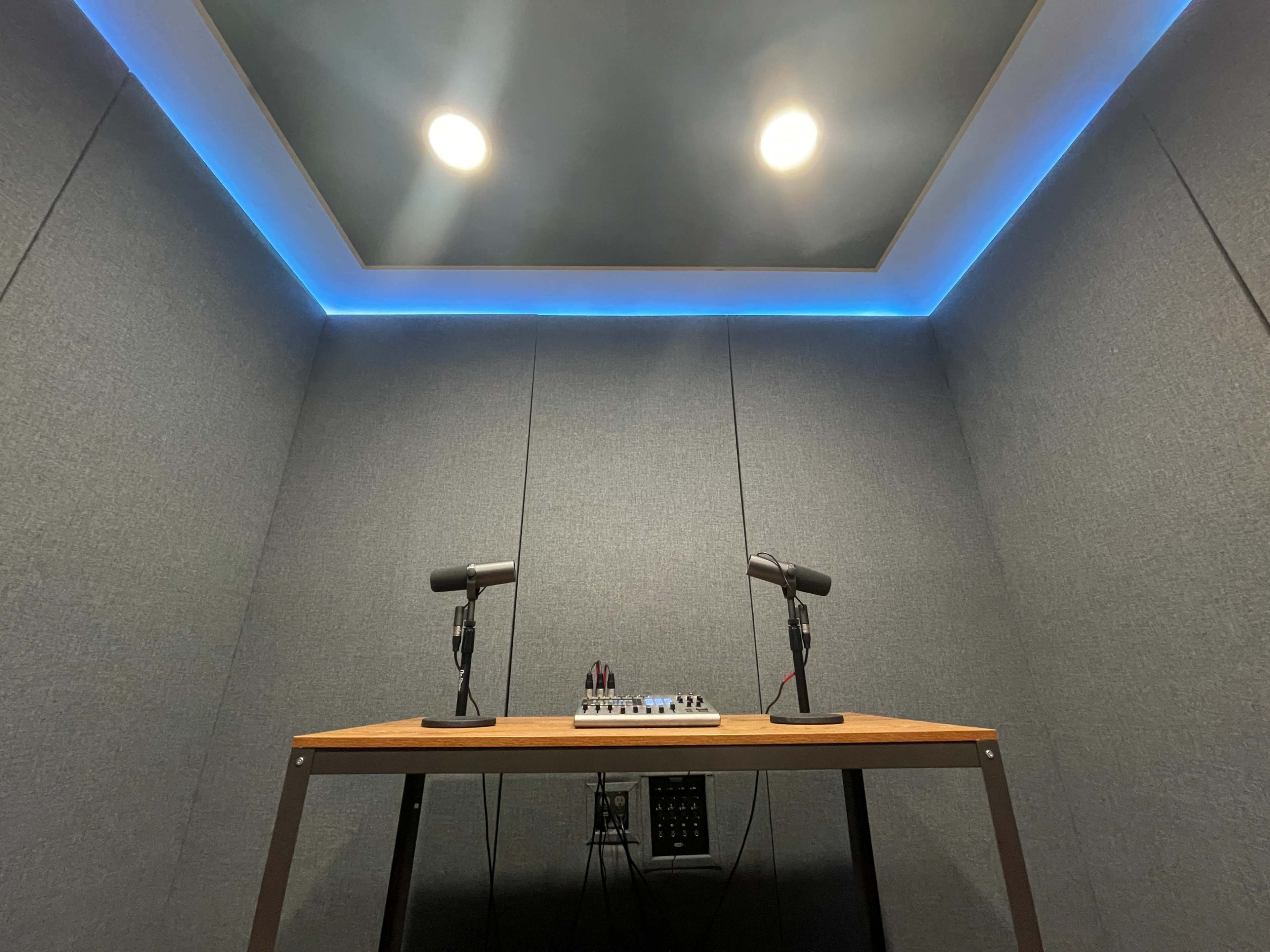 A soundproof recording studio featuring a wooden table with two microphones and an audio mixer beneath a lit ceiling.
