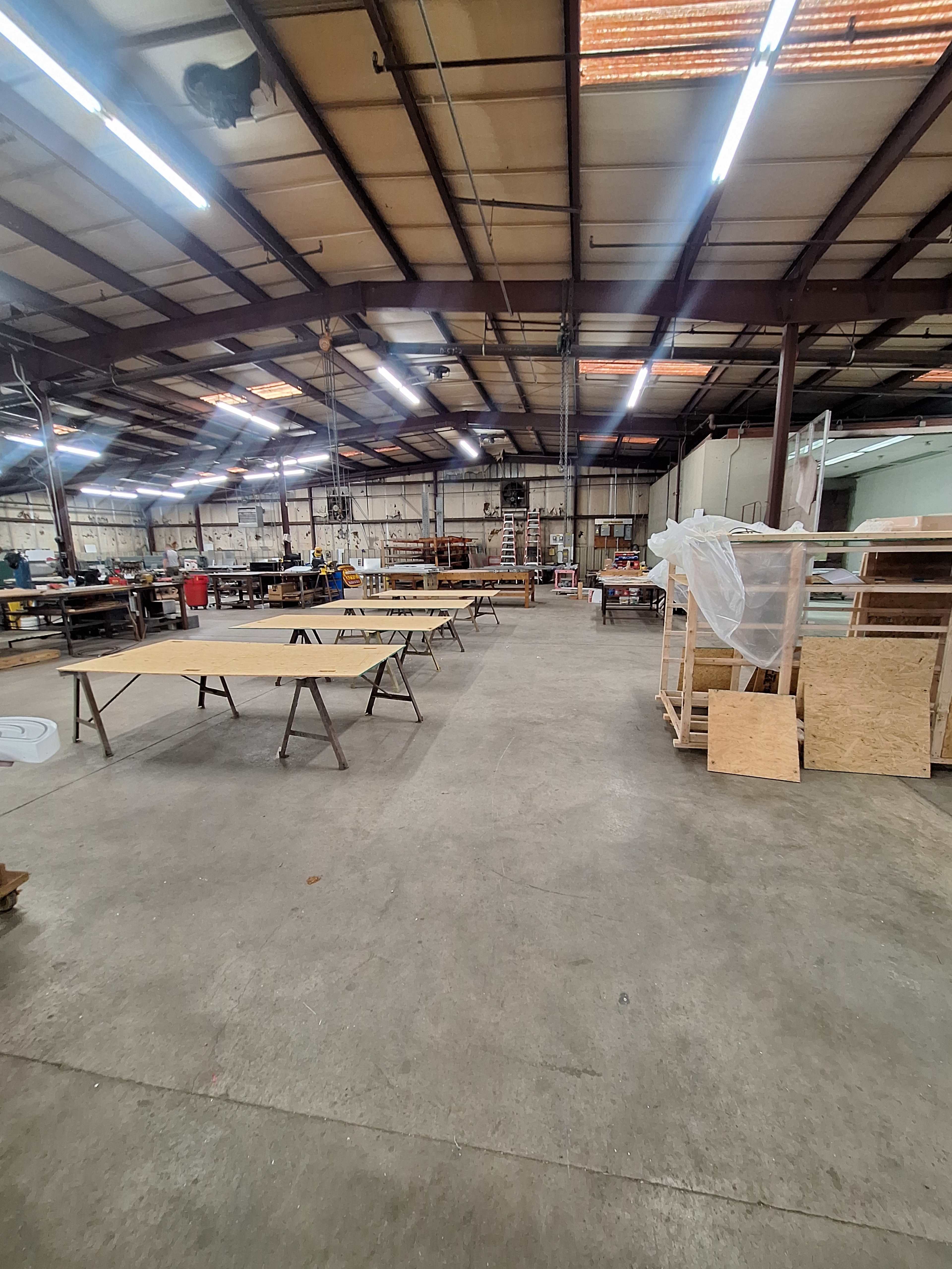 The image shows a spacious industrial workshop with several worktables arranged throughout and various materials and tools scattered around.