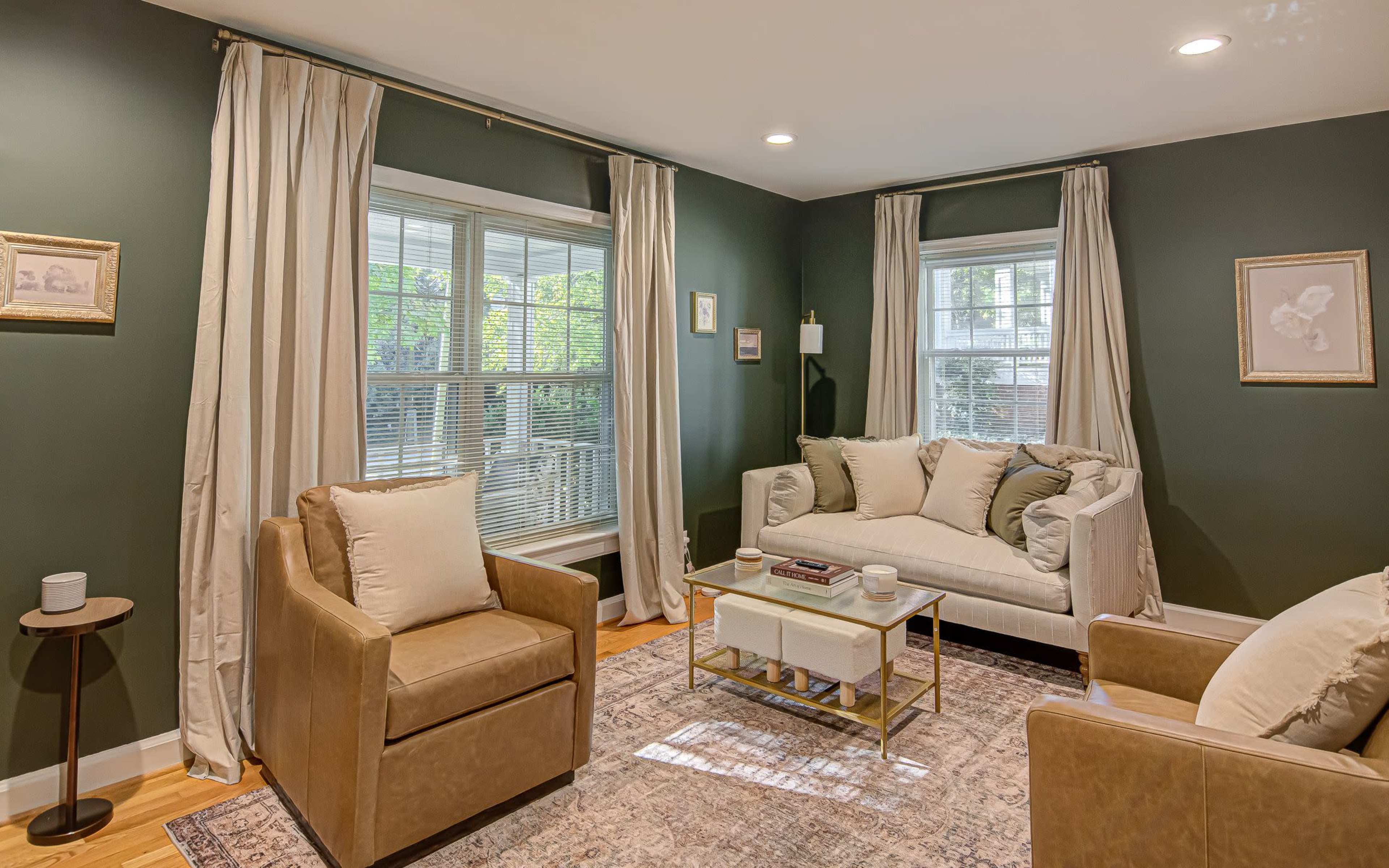 The living room features a white sofa and two tan chairs, arranged around a gold coffee table, with large windows and muted green walls.