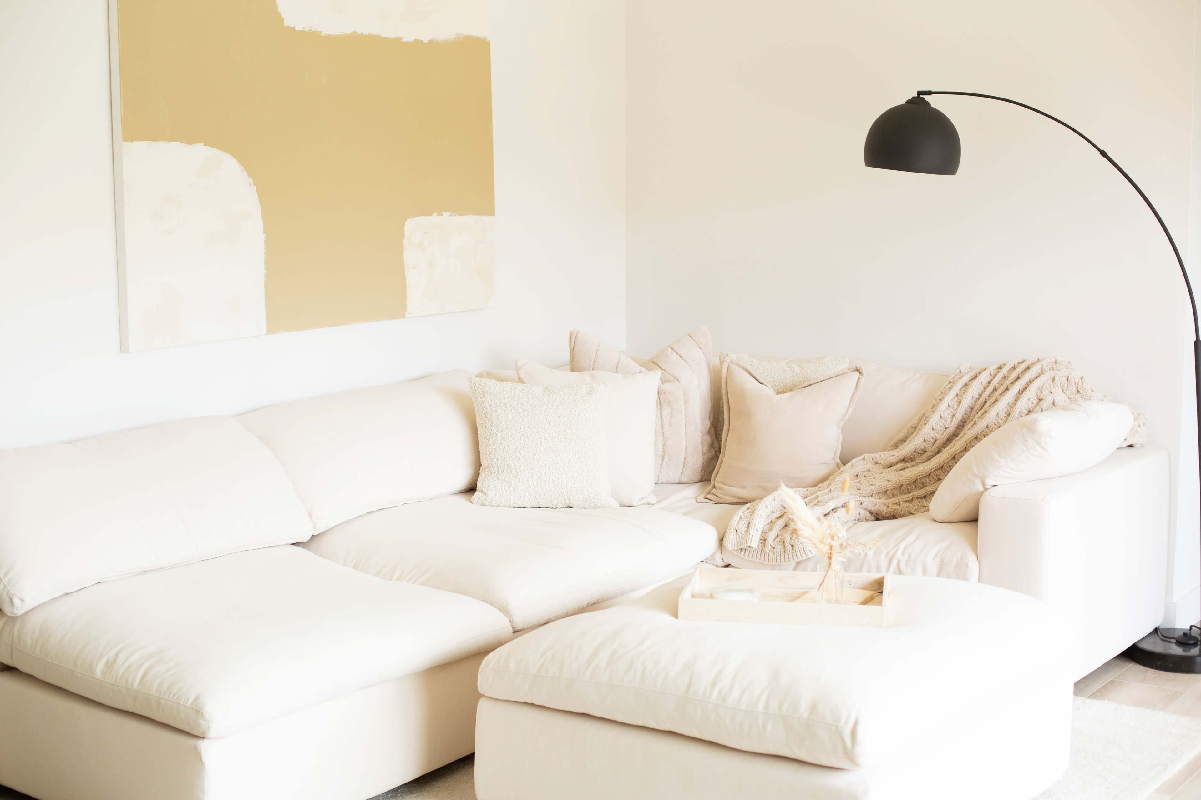 A light-colored sectional sofa in a minimalist living room, accompanied by a floor lamp and a painting on the wall.