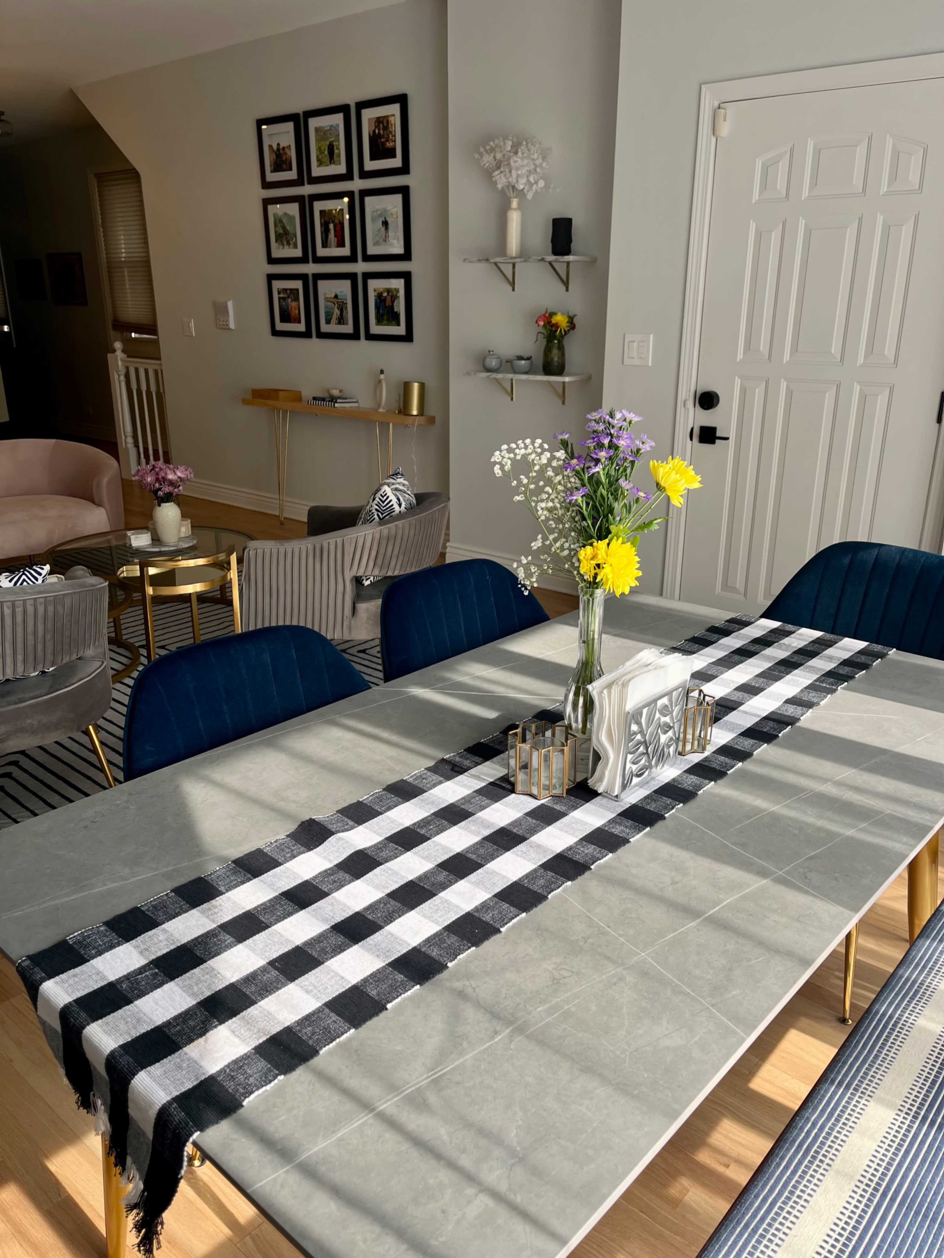 A dining table set with a black and white checked runner is adorned with a vase of flowers, against a backdrop of framed photos on the wall and a cozy living area.