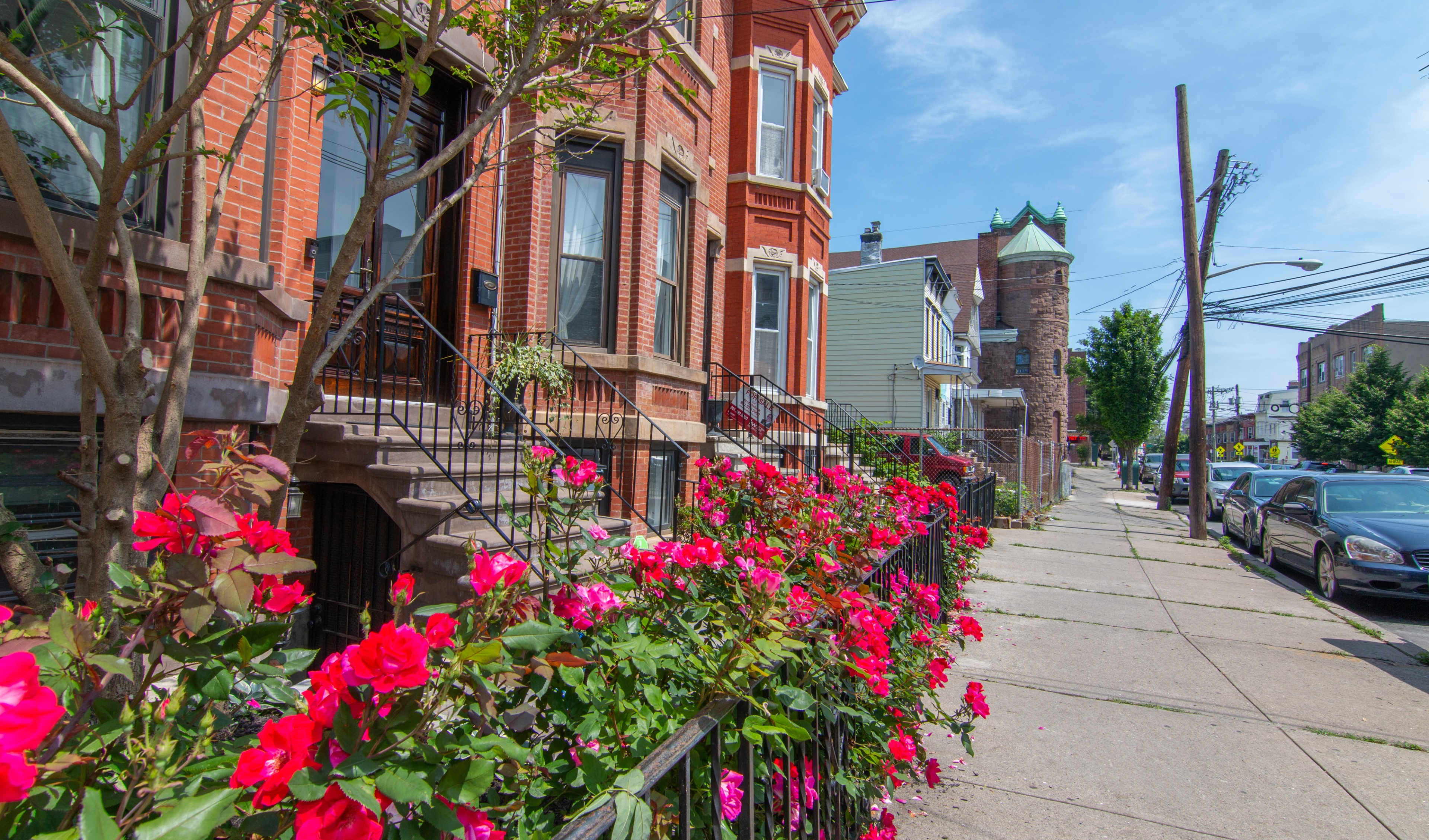 A row of brick buildings lines a sidewalk adorned with vibrant pink flowers and a clear blue sky above.
