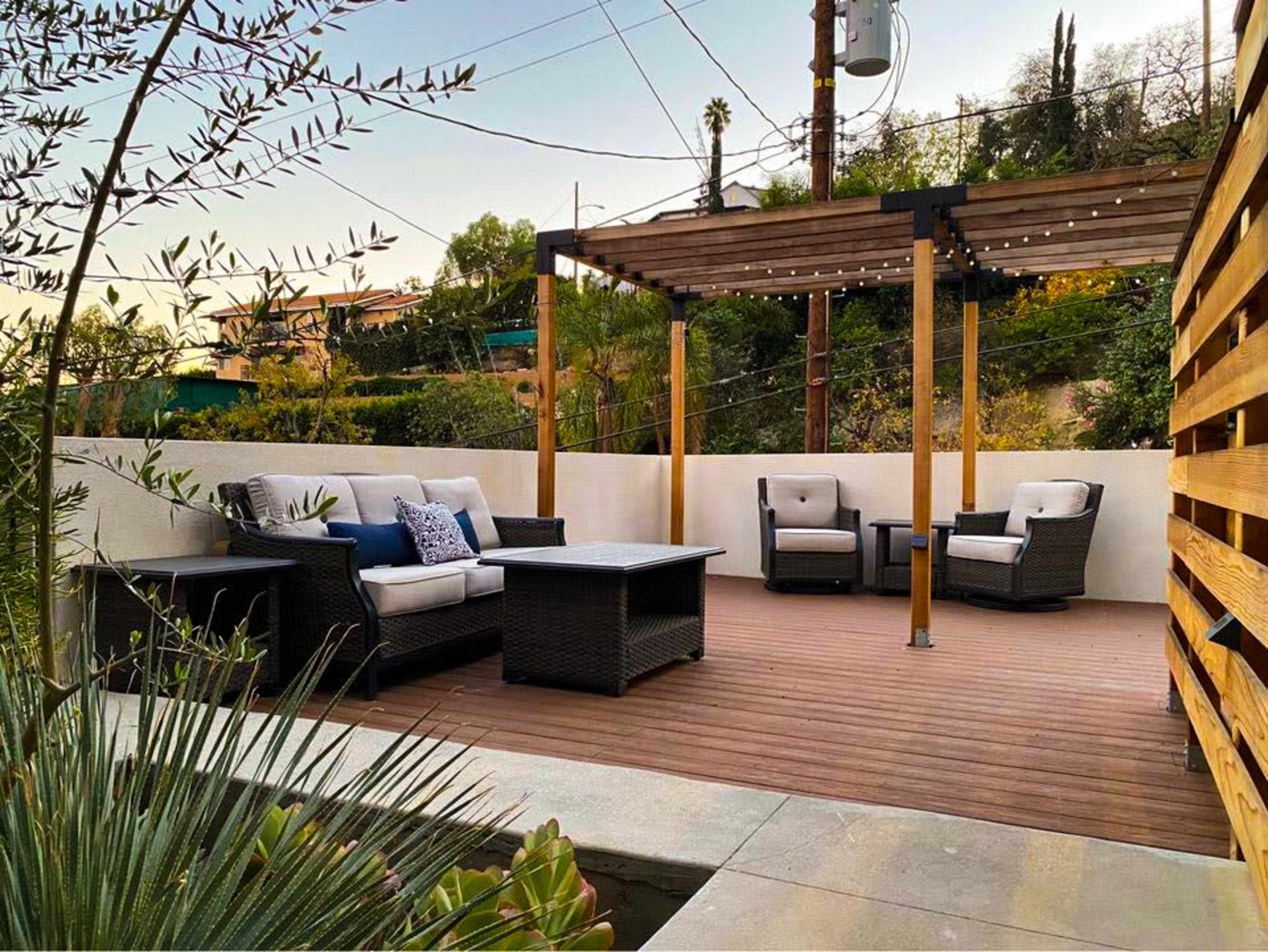 A wooden deck with outdoor seating and a pergola is positioned in a landscaped area surrounded by greenery.