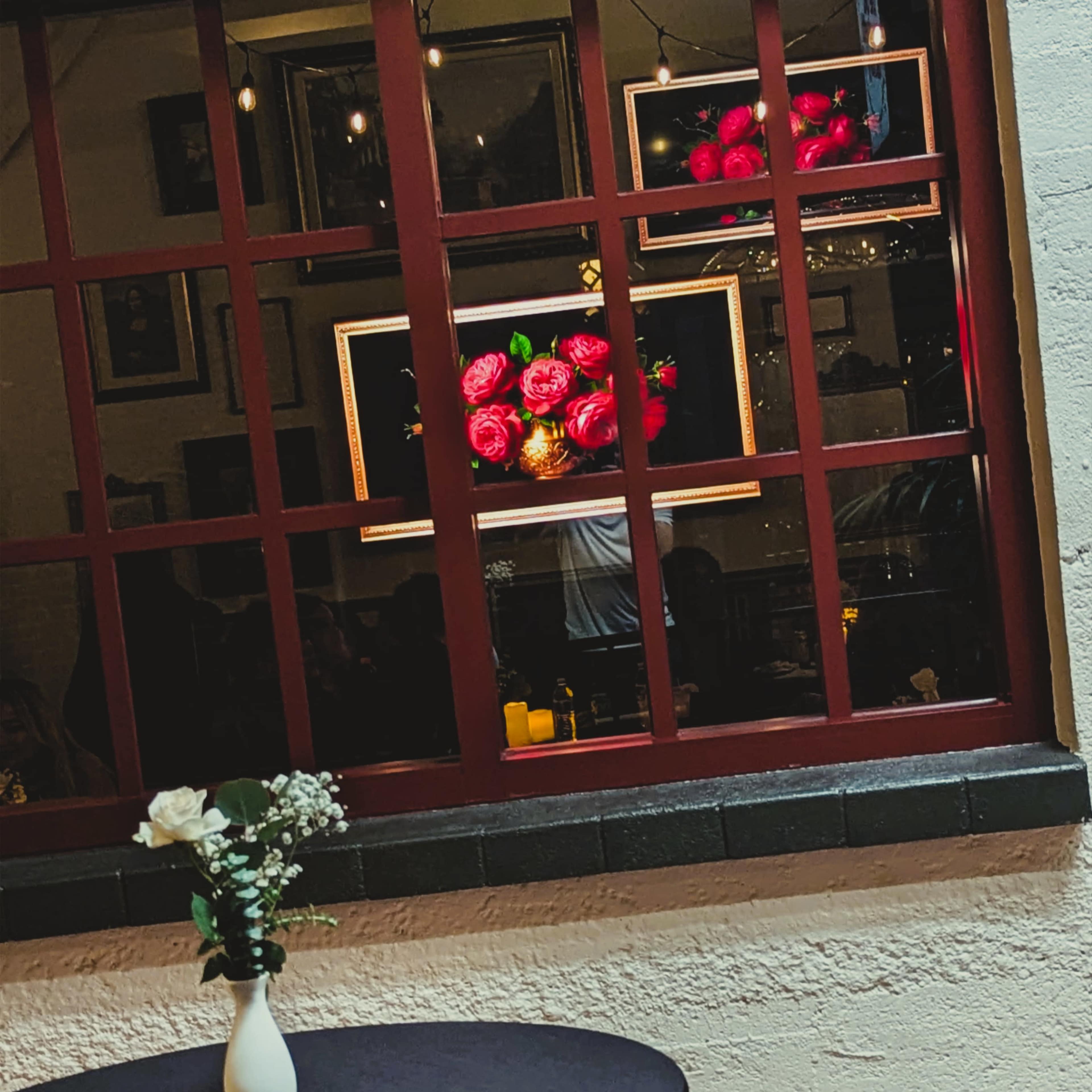 A table with a vase of flowers sits in front of a window displaying illuminated floral artwork and a dimly lit interior.