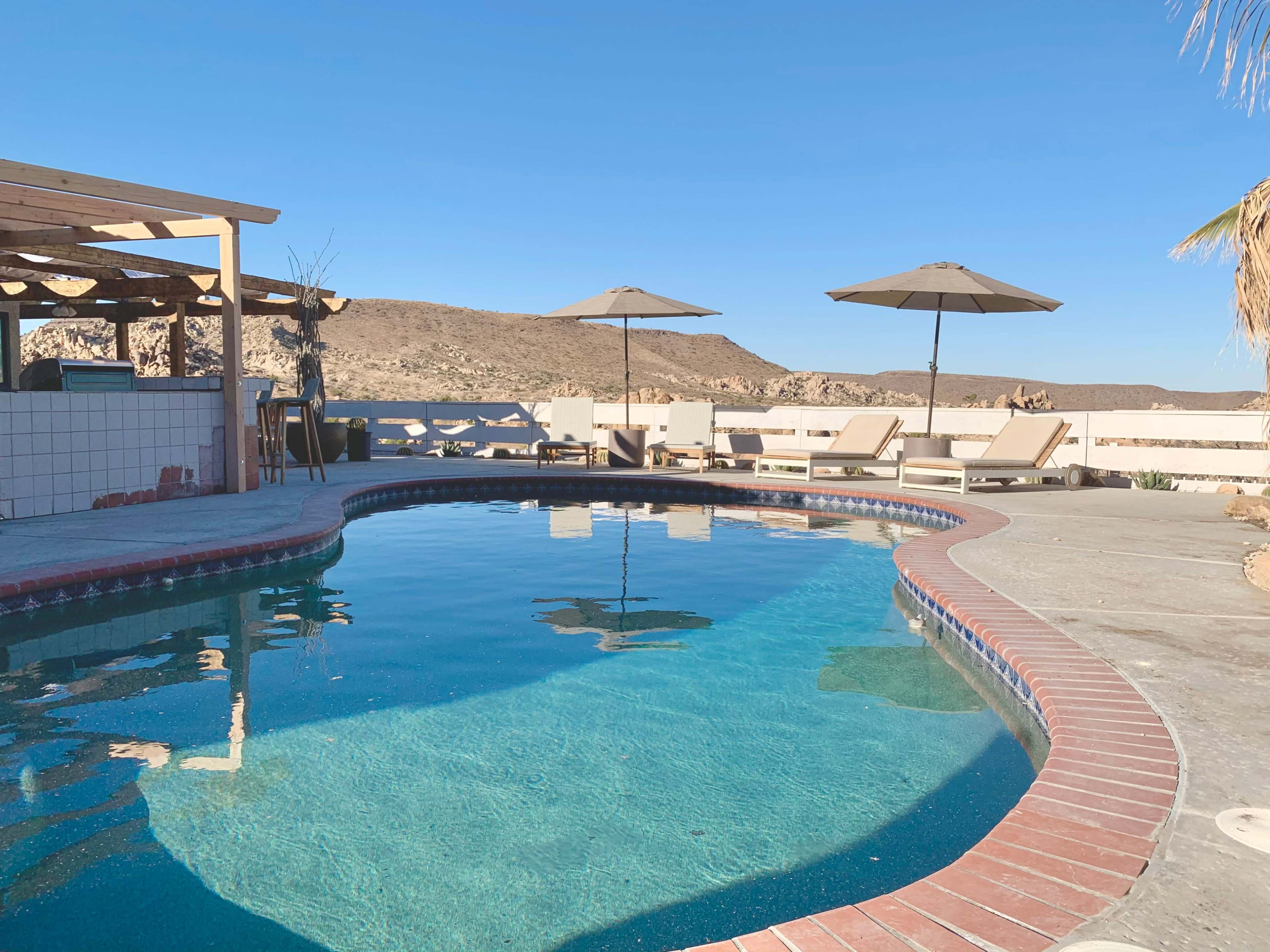 A swimming pool with lounge chairs and umbrellas is set against a mountainous desert landscape.