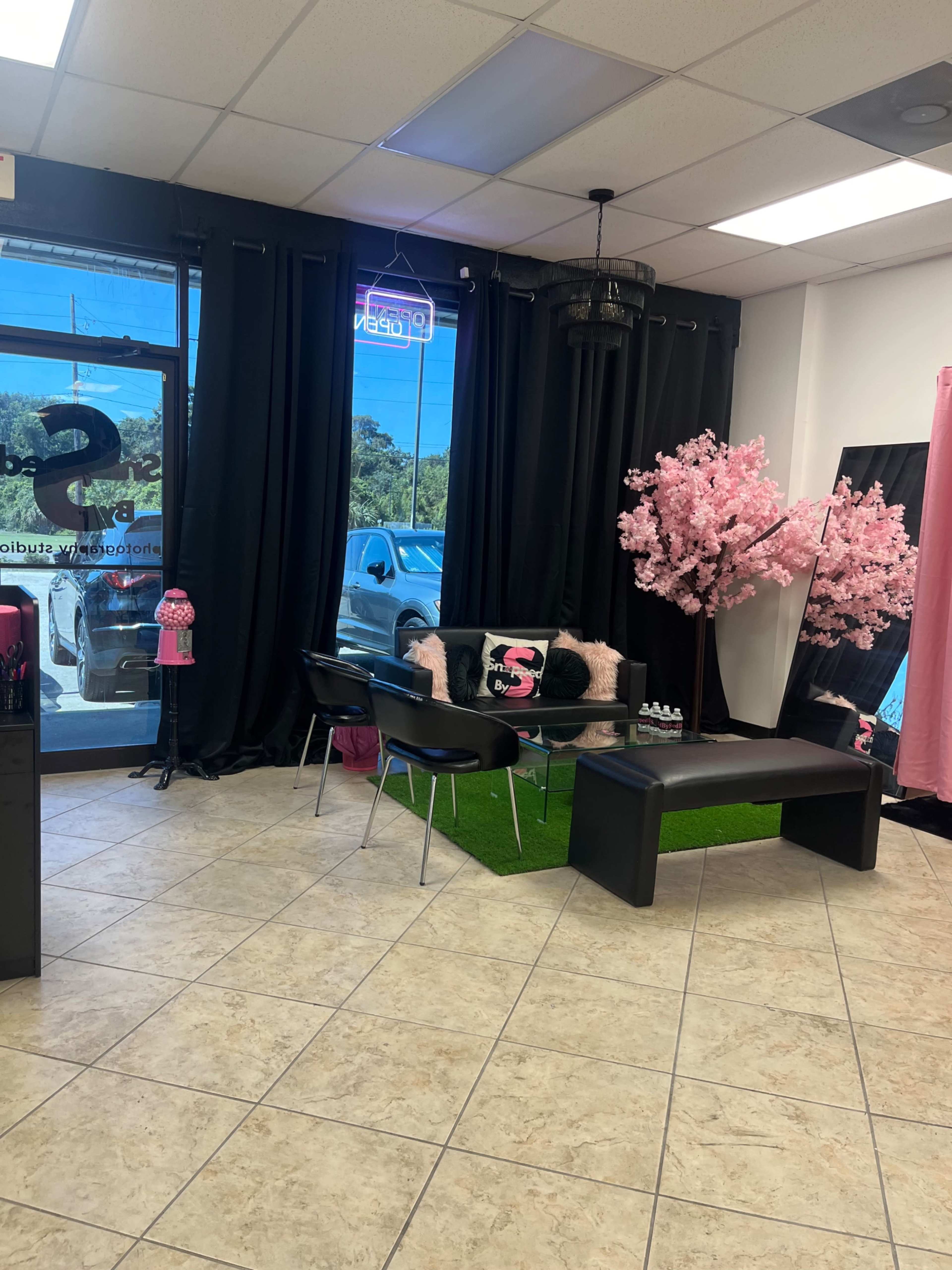 The image shows a salon interior with a seating area featuring black chairs, a low table, a faux grass rug, and a decorative pink cherry blossom tree under a chandelier.