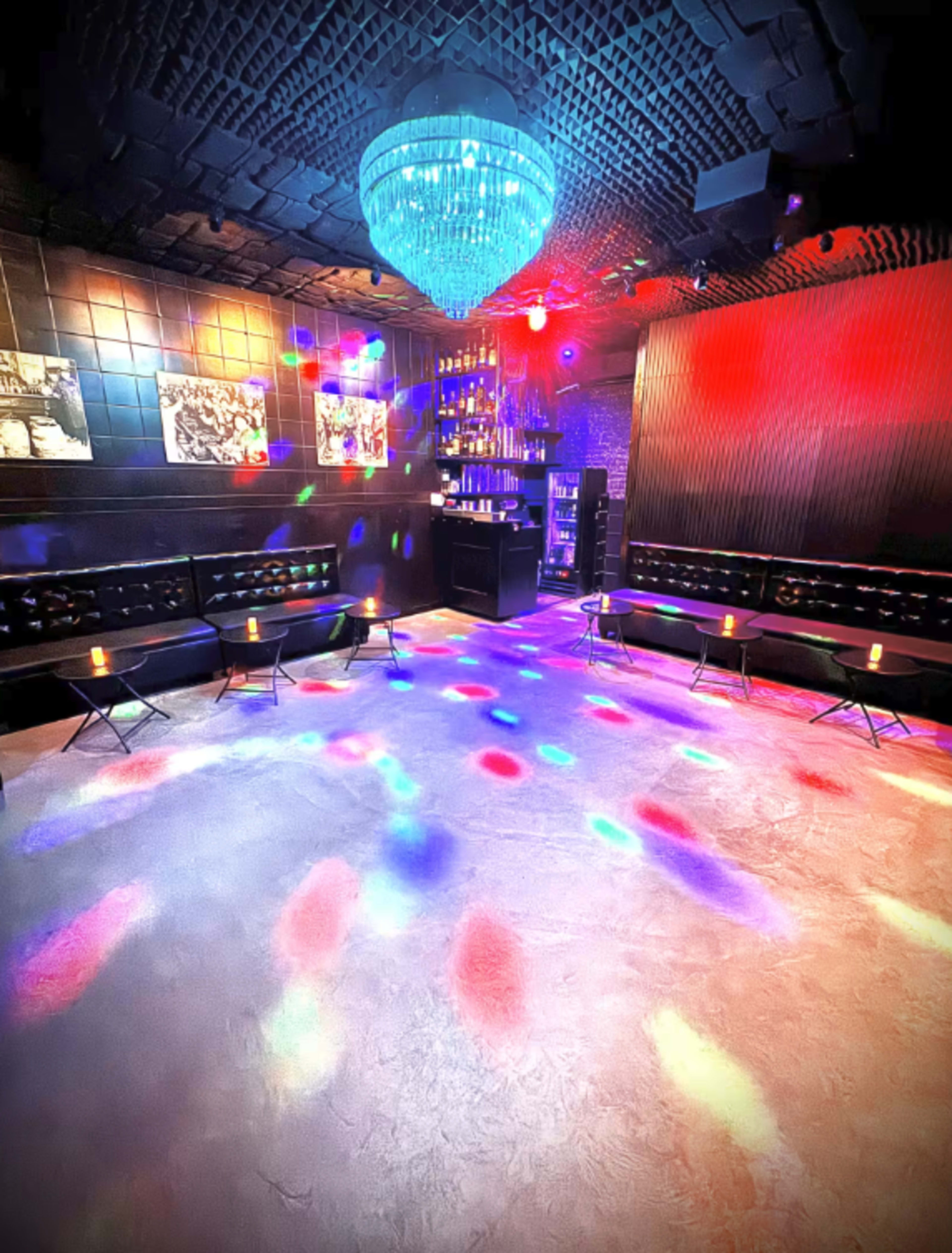 The image shows a dimly lit lounge with modern furnishings, a bar area, and colorful lights reflecting on the floor.