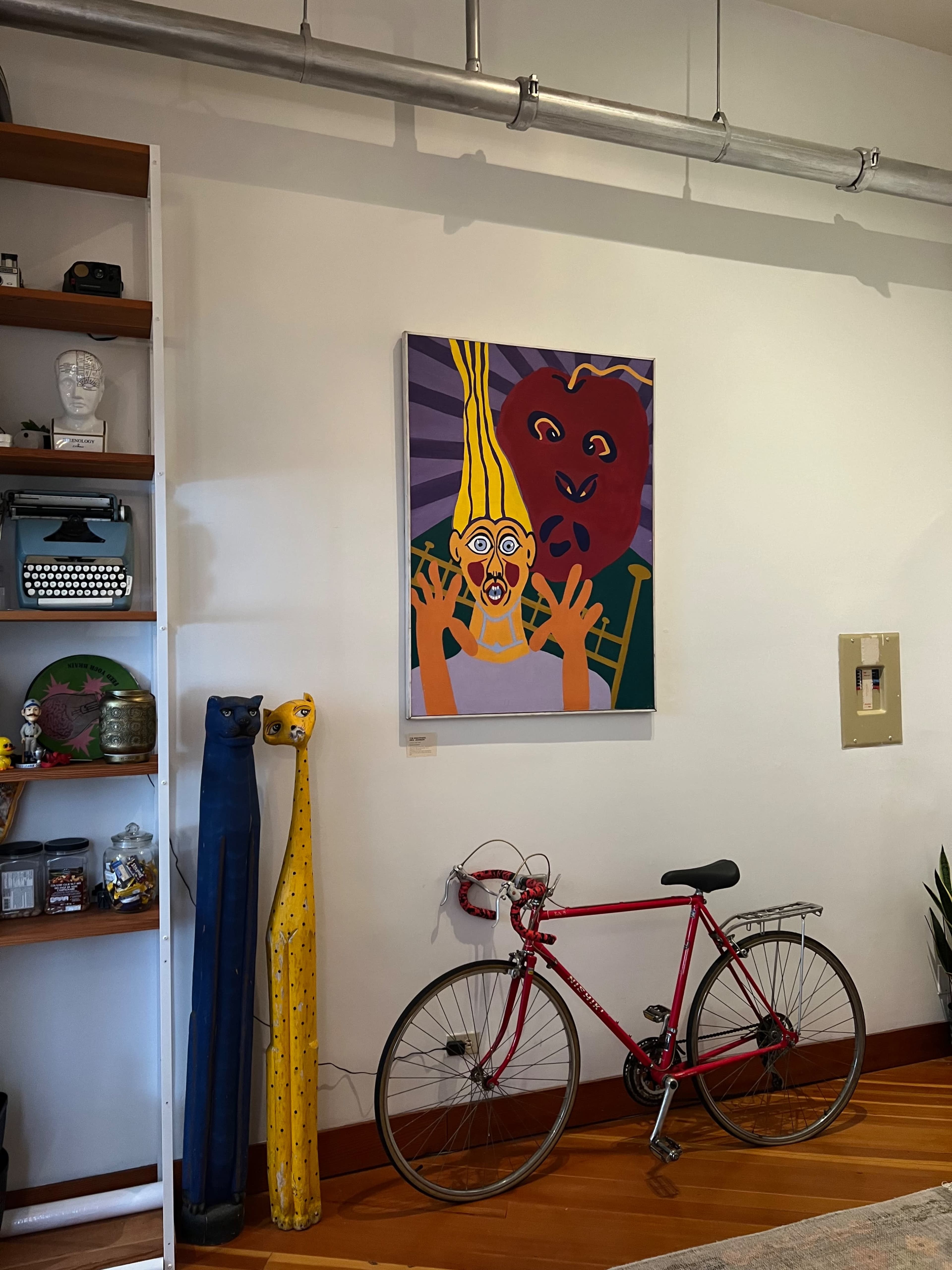 A colorful painting of abstract faces on a wall, with a pink bicycle leaned against it and a shelf displaying various items nearby.